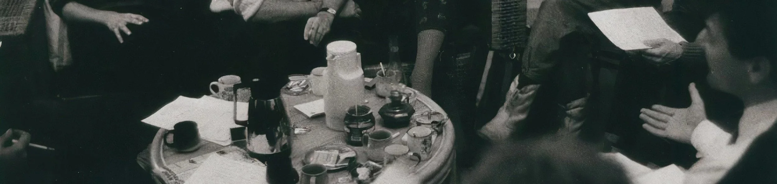 People gathered around a coffee table with cups, mugs, bottles, and snacks, engaging in conversation and discussion.