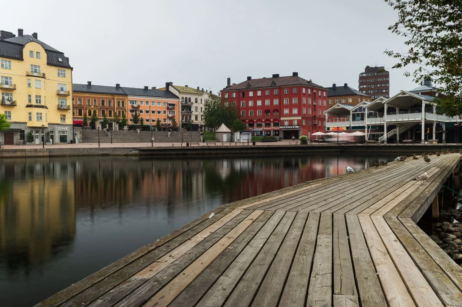 View of colorful buildings along a waterfront with their reflections in the water, wooden dock in the foreground, and overcast sky. Södertälje