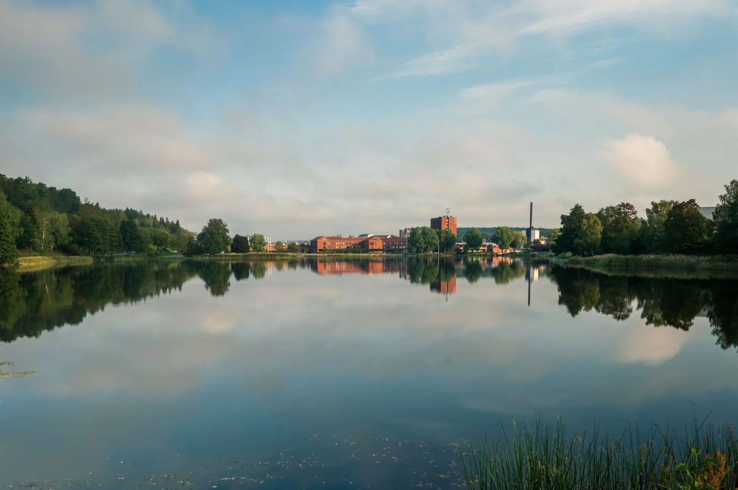 A calm river reflecting trees on both banks and buildings in the distance, with a partly cloudy sky overhead. Södertälje