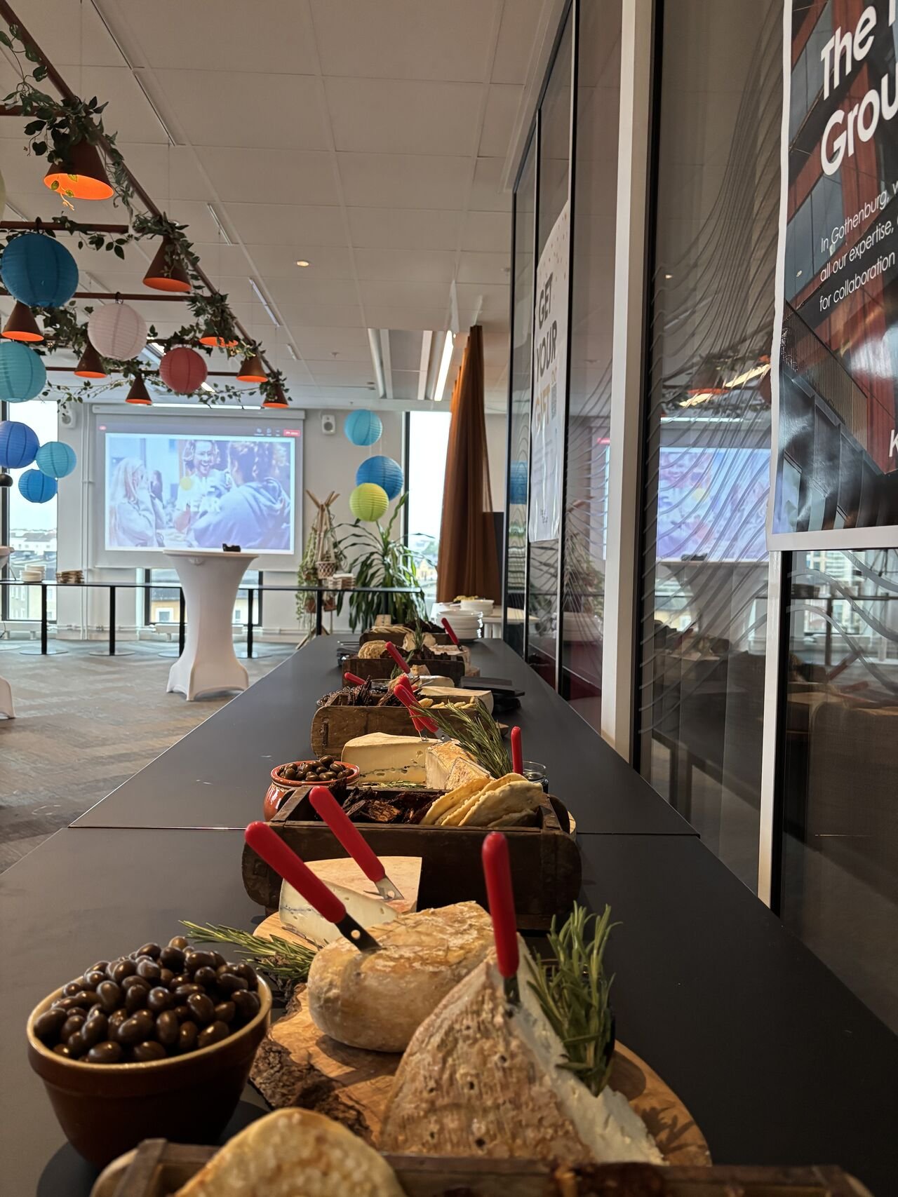A table with cheese, bread, olives, and garnishes at a party with colorful paper lanterns and a projector screen in the background.