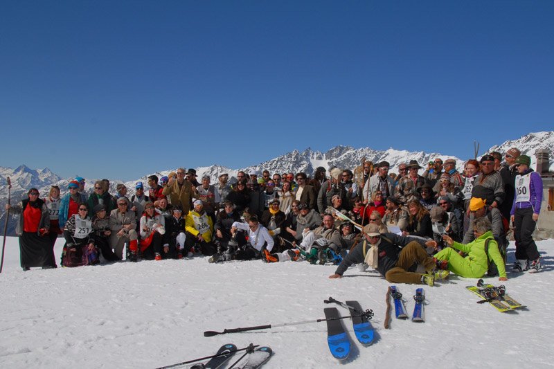 Group of tourists and skiers on a snowy mountainside with snow-capped peaks in the background, some posing for the photo while others prepare to ski.