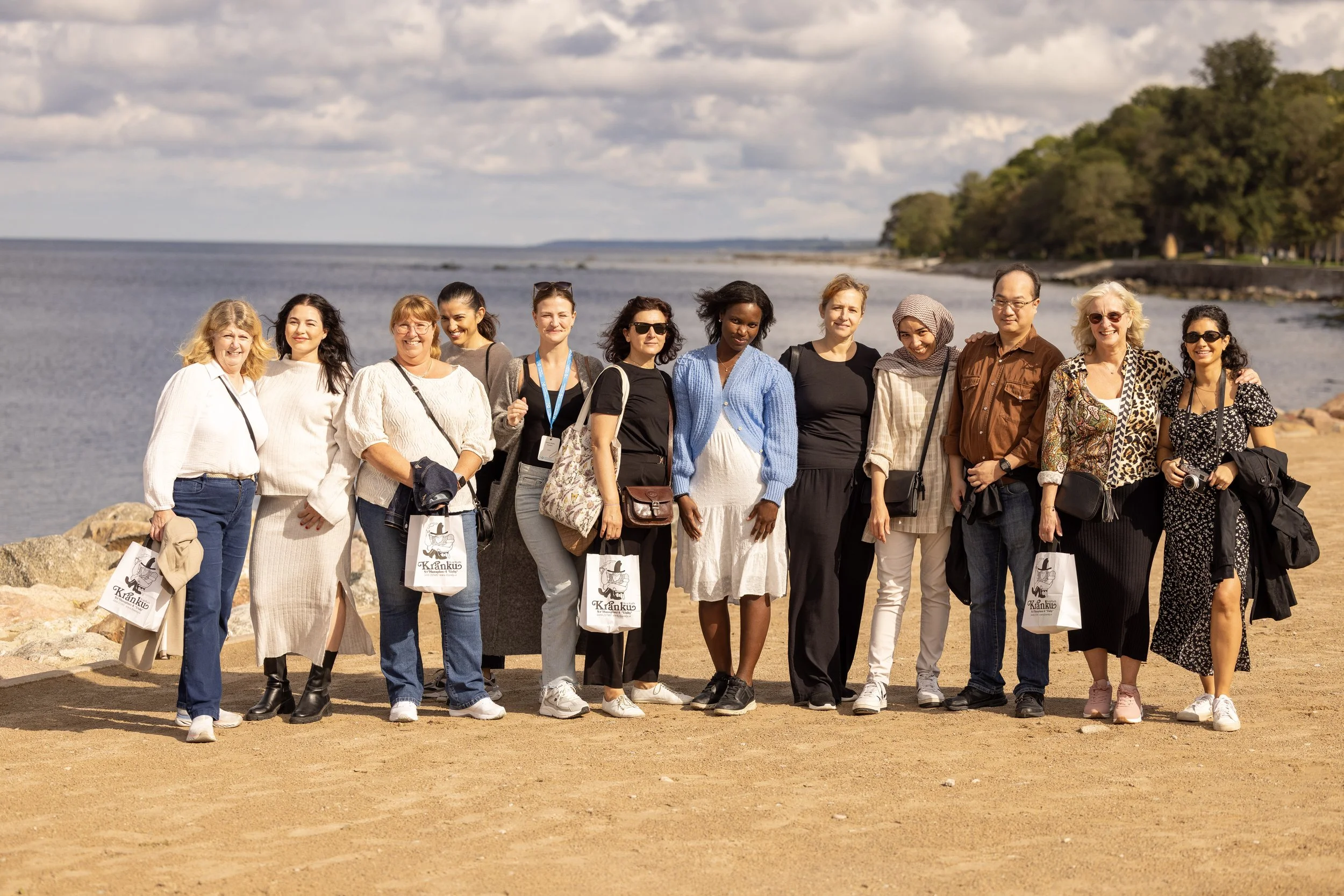 Group of twelve women and one man standing on a sandy beach on gotland visby near water with trees in the background.