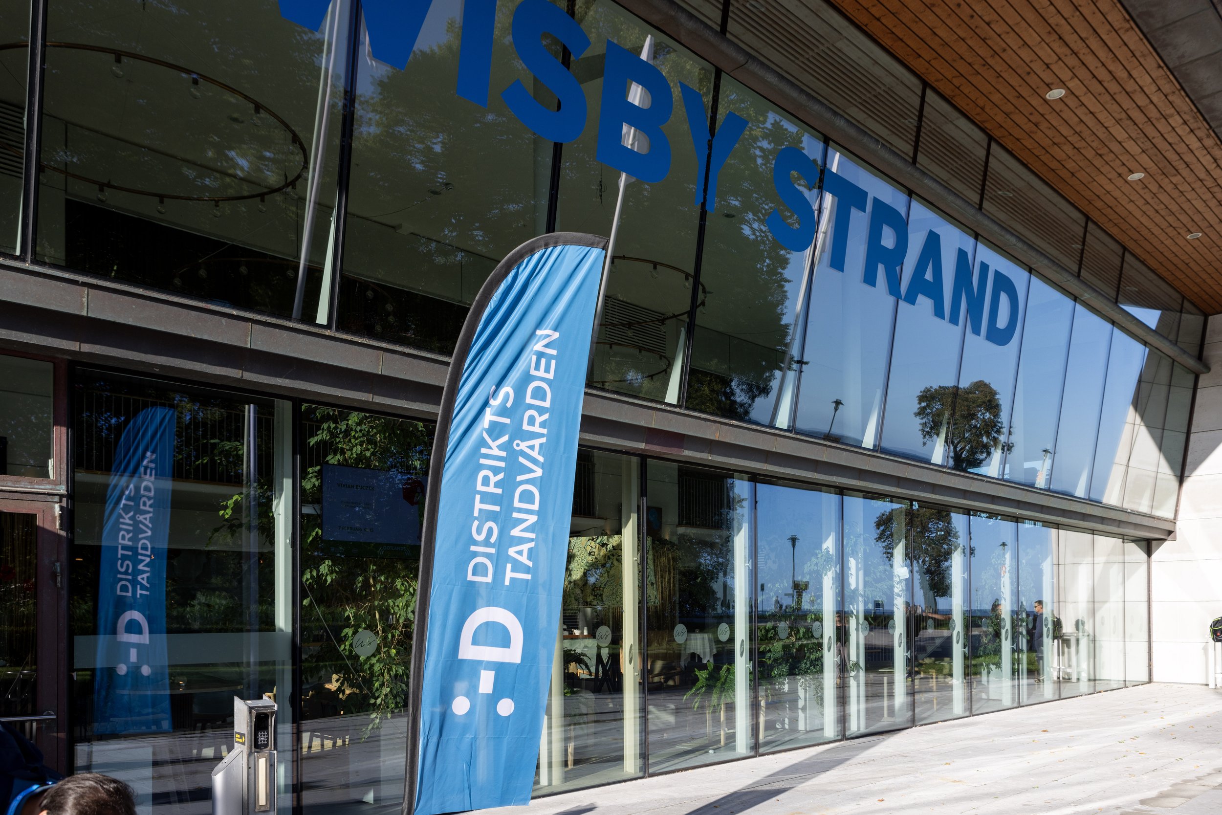 Exterior view of a modern building with large glass windows and wooden ceiling wisby strand, with blue banners and letters that read 'Distrikstandvården'