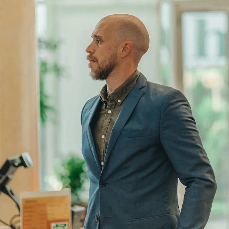 A man with a beard and shaved head wearing a blue suit jacket and a dark shirt, standing indoors by a window, looking to the side with a serious expression.