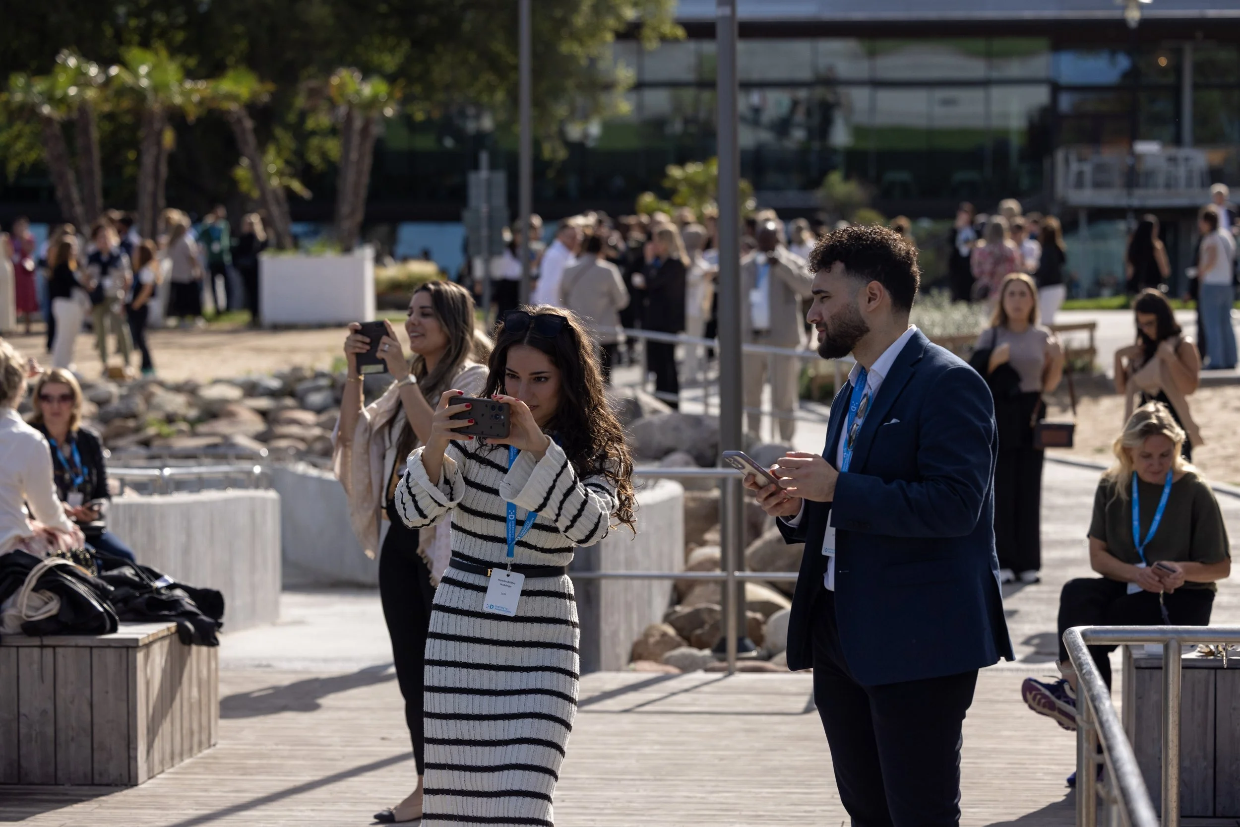 People taking photos and looking at their phones outdoors at a sunny event with many attendees in the background.