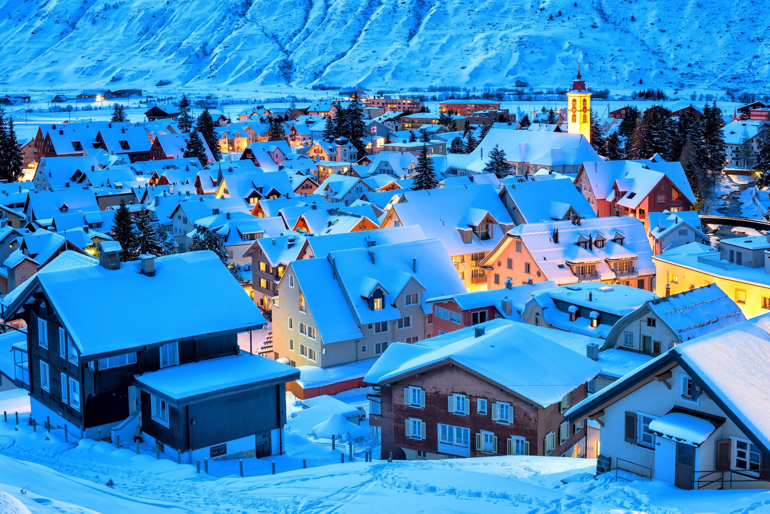 Snow-covered houses and buildings in a mountain town at dusk, with illuminated windows and a backdrop of snow-covered mountains.