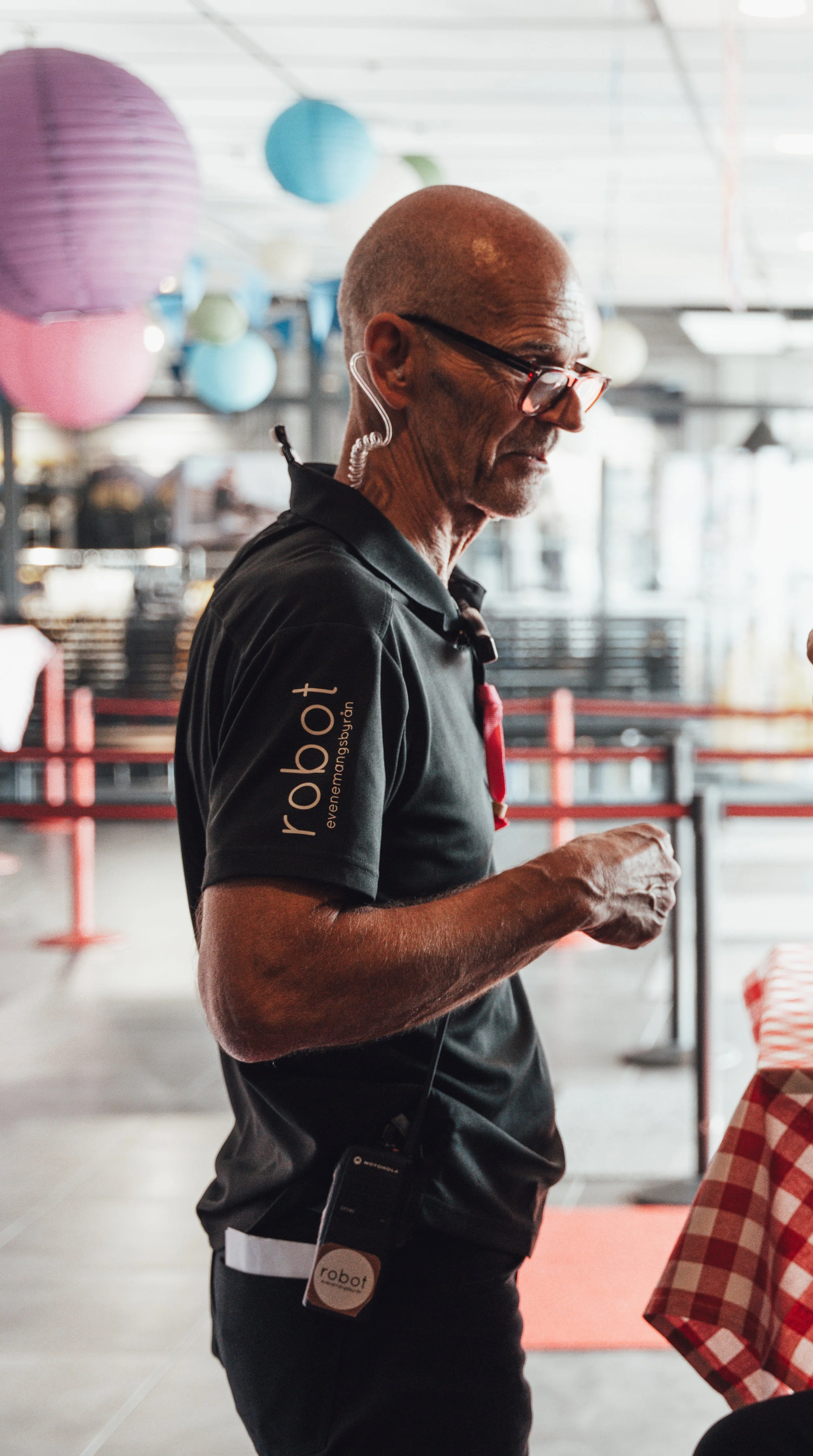 A man wearing a black shirt with the word "robot" and a logo on the sleeve, standing in a dining area with colorful hanging lanterns, using a smartphone.