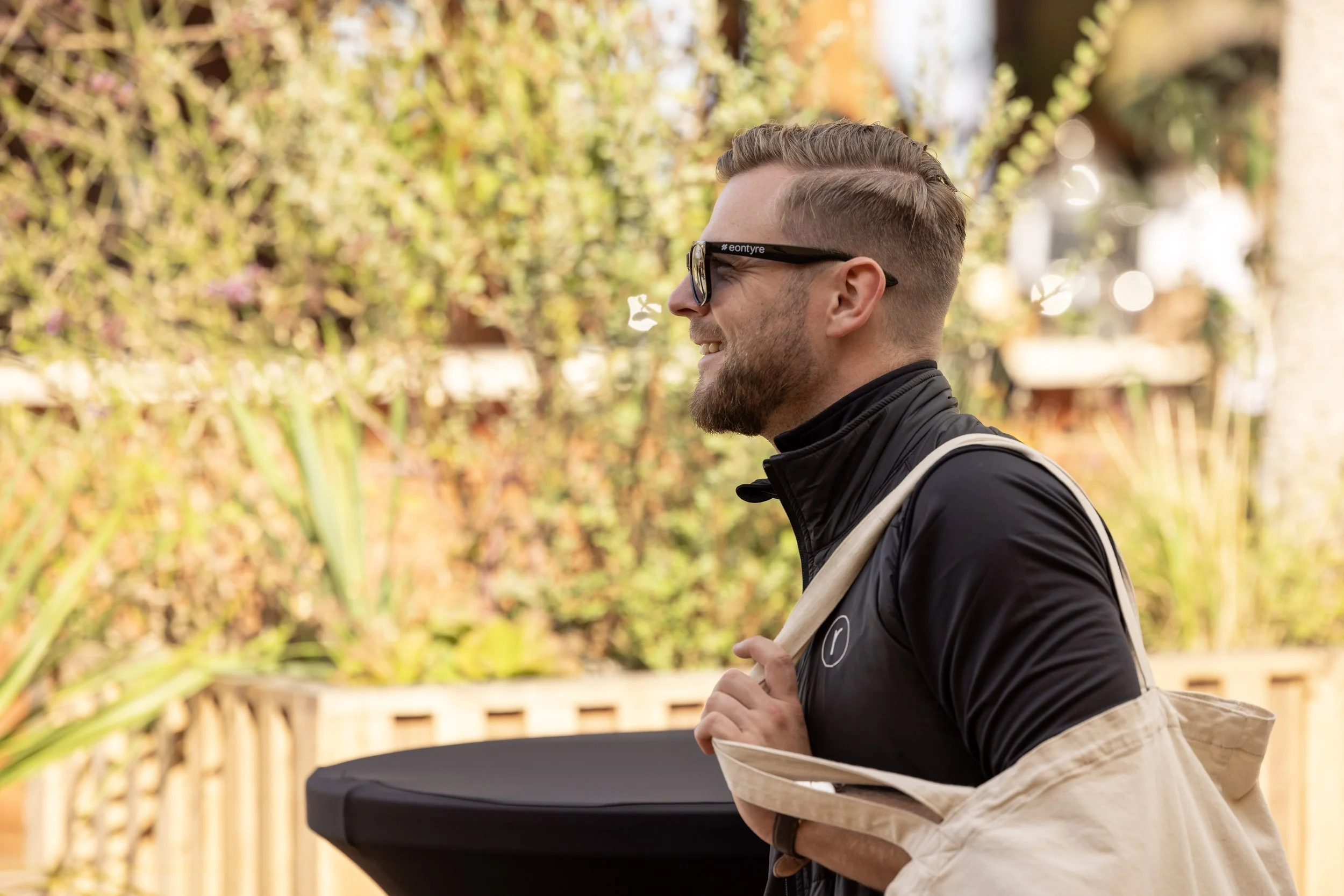 A man with short hair and a beard, wearing glasses and a black jacket, smiling while holding a tote bag over his shoulder, outdoors with a background of plants and a wooden fence.