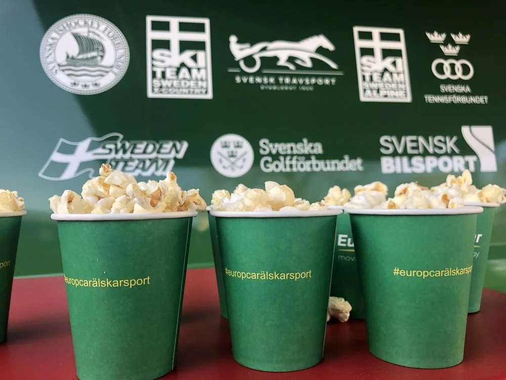 Green cups filled with popcorn in front of a backdrop with Swedish sports logos and text.