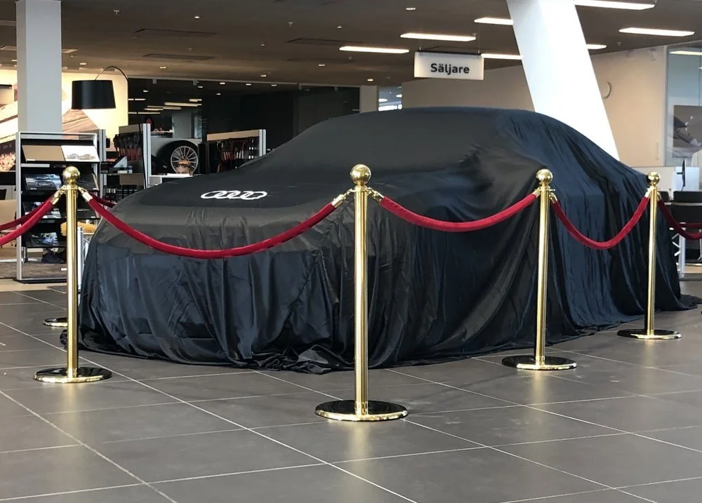 New Audi car covered with black fabric in a showroom, surrounded by red velvet ropes and gold stanchions.