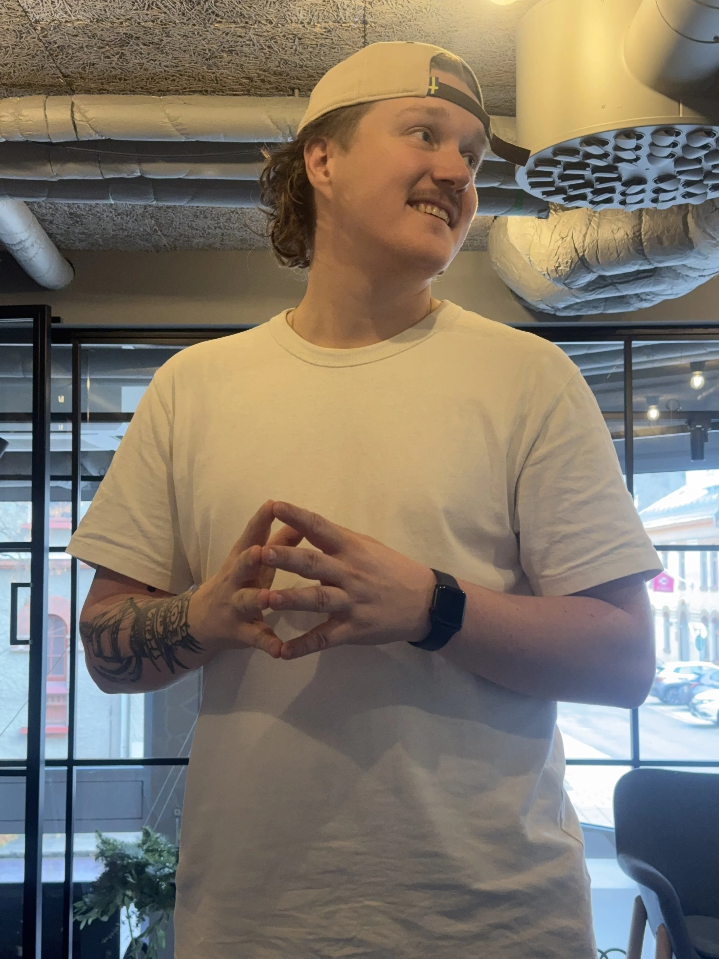 A young man with curly hair wearing a white t-shirt, a backwards baseball cap, and a smartwatch, smiling and standing indoors near large windows and ceiling ductwork.