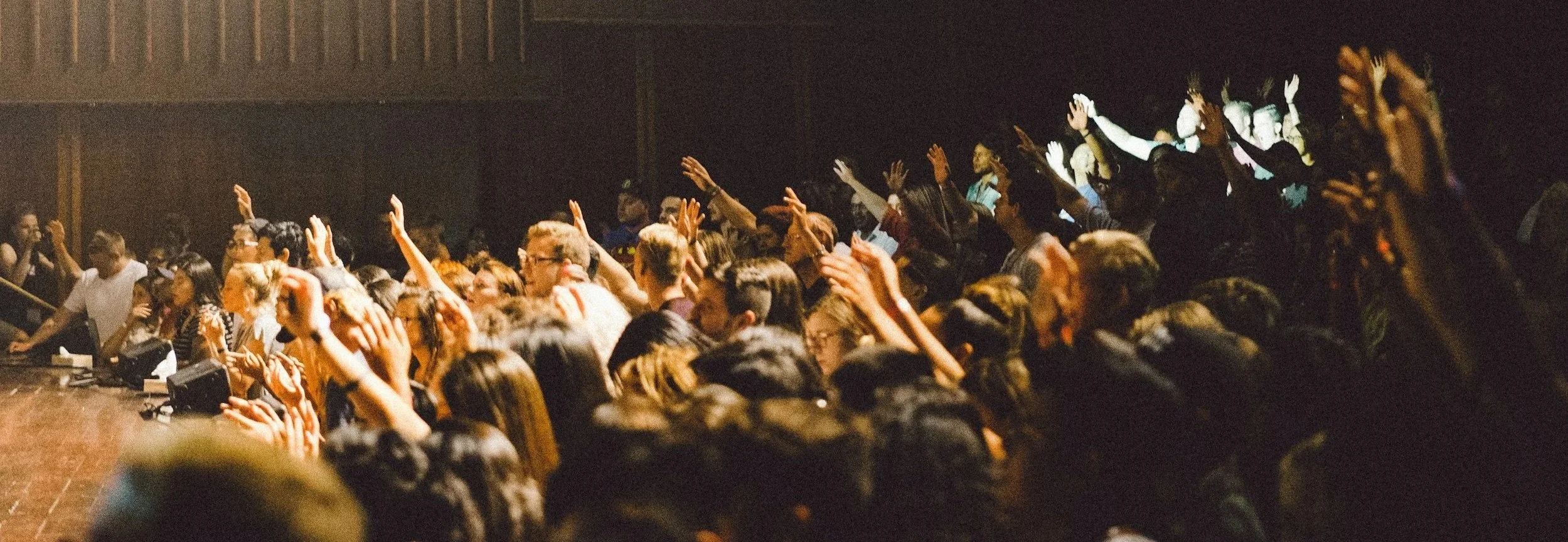 Audience seated in a dark theater, raising their hands during a performance or presentation.