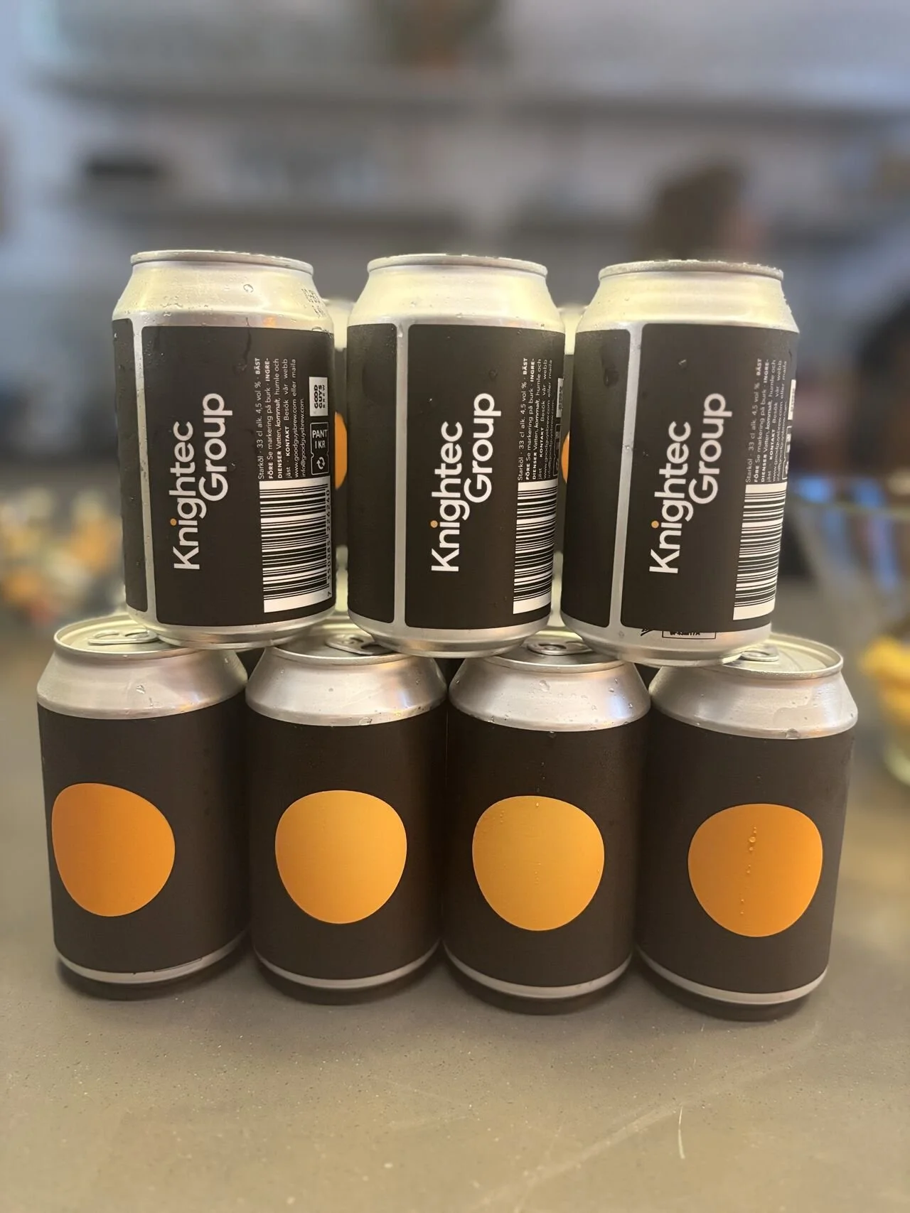 Six cans stacked in two layers, with three cans on the bottom and three cans on the top. The cans have black labels with orange circles and white text that reads 'Knightc Group.'