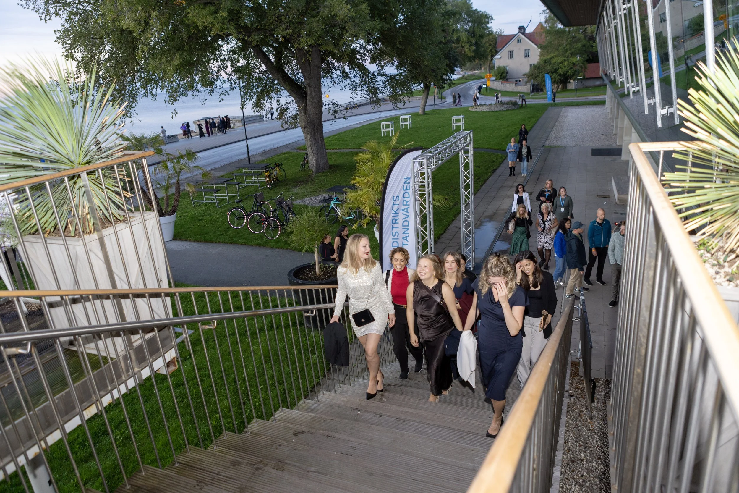 A group of people ascending outdoor stairs near a river in visby, with greenery and bicycles nearby and a large tree in the background during daytime.