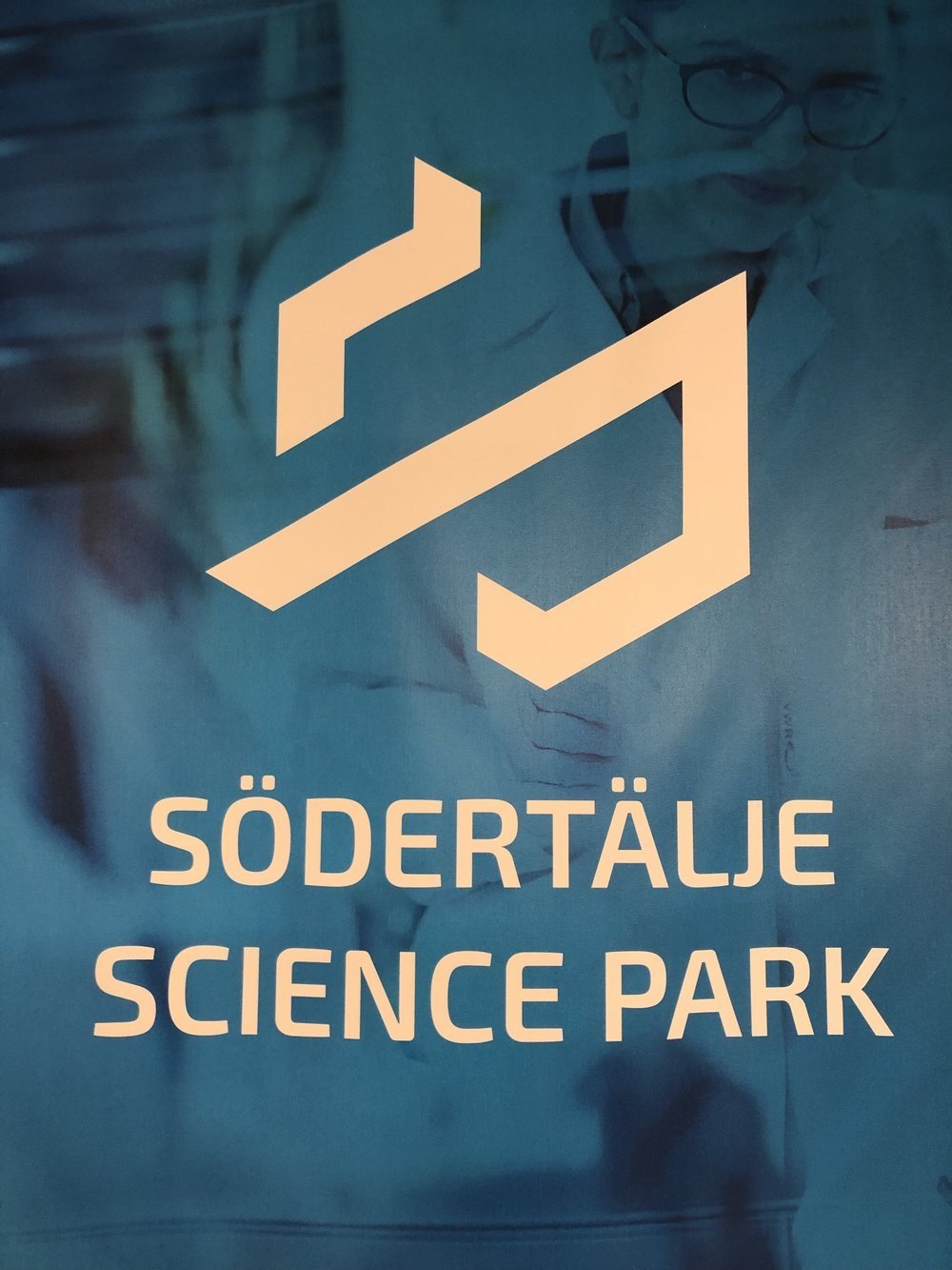 Sign for Södertälje Science Park with a logo, and a background image of a curious woman with glasses examining a test tube in a laboratory.