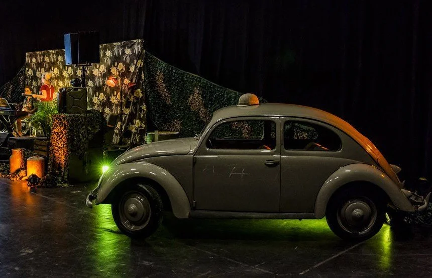 Vintage gray Volkswagen Beetle on stage with a band playing in the background.