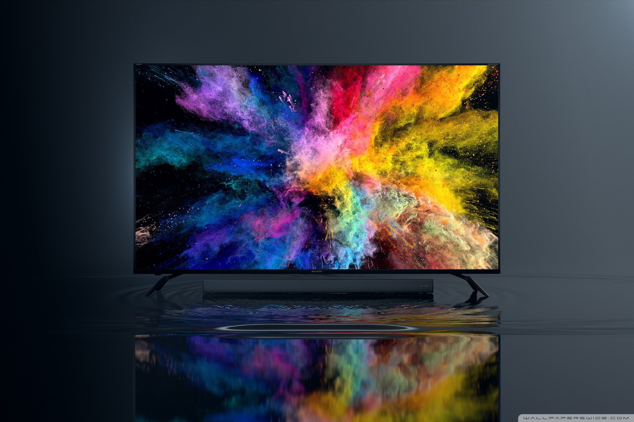 A flat-screen television displaying a vibrant explosion of multicolored powder or paint against a dark background, with the image reflected on a glossy surface below.