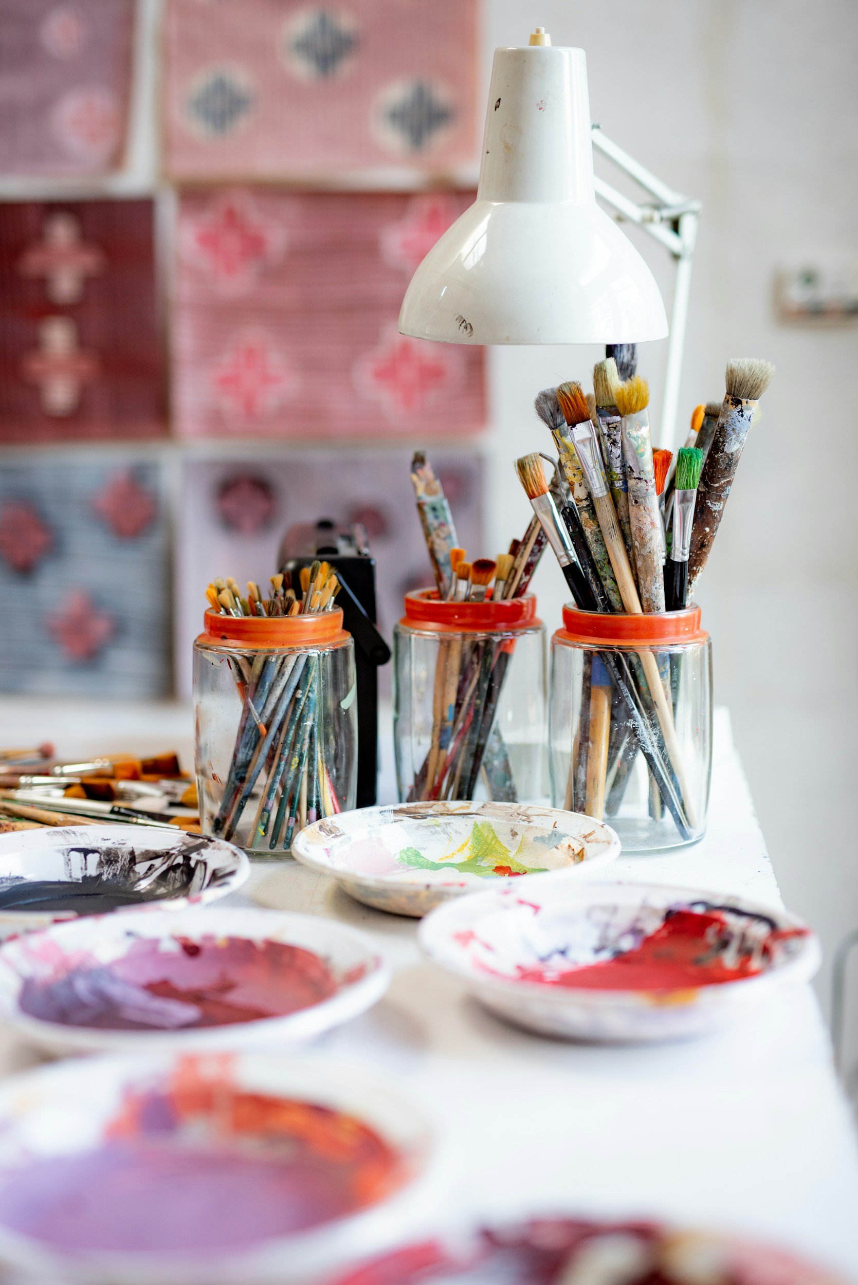 An artist's workspace with paintbrushes in jars and plates of mixed paints on a table, with a lamp overhead and colorful artwork in the background.