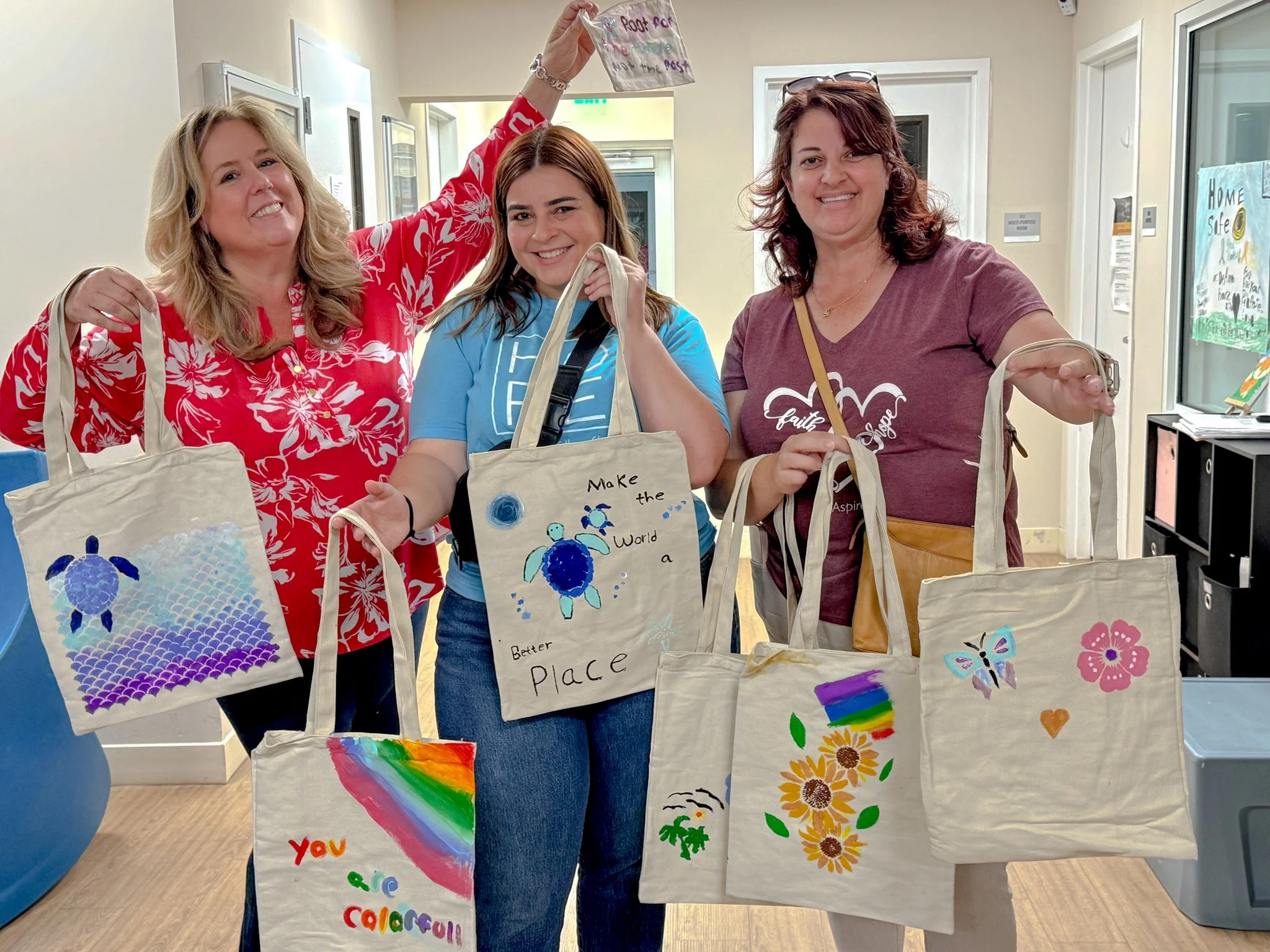 Three women are standing indoors, holding off-white tote bags painted with colorful designs. The woman on the left has blond hair and is wearing a red floral shirt, the woman in the middle has brown hair and is wearing a blue shirt, and the woman on the right has curly hair and is wearing a maroon shirt. All three women are smiling and showing their bags, which feature messages like 'You are colorful' and 'Make the world a better place,' along with various painted images of animals, rainbows, and flowers.