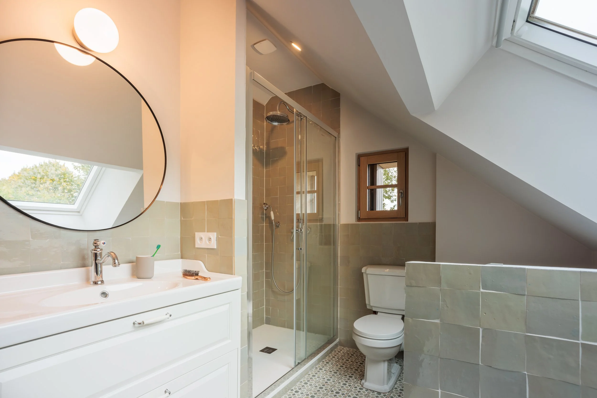 Salle de bain avec lavabo blanc, miroir rond, douche en verre, toilette, petites fenêtres en bois, plafond en pente, lumière naturelle depuis un toit en verre.