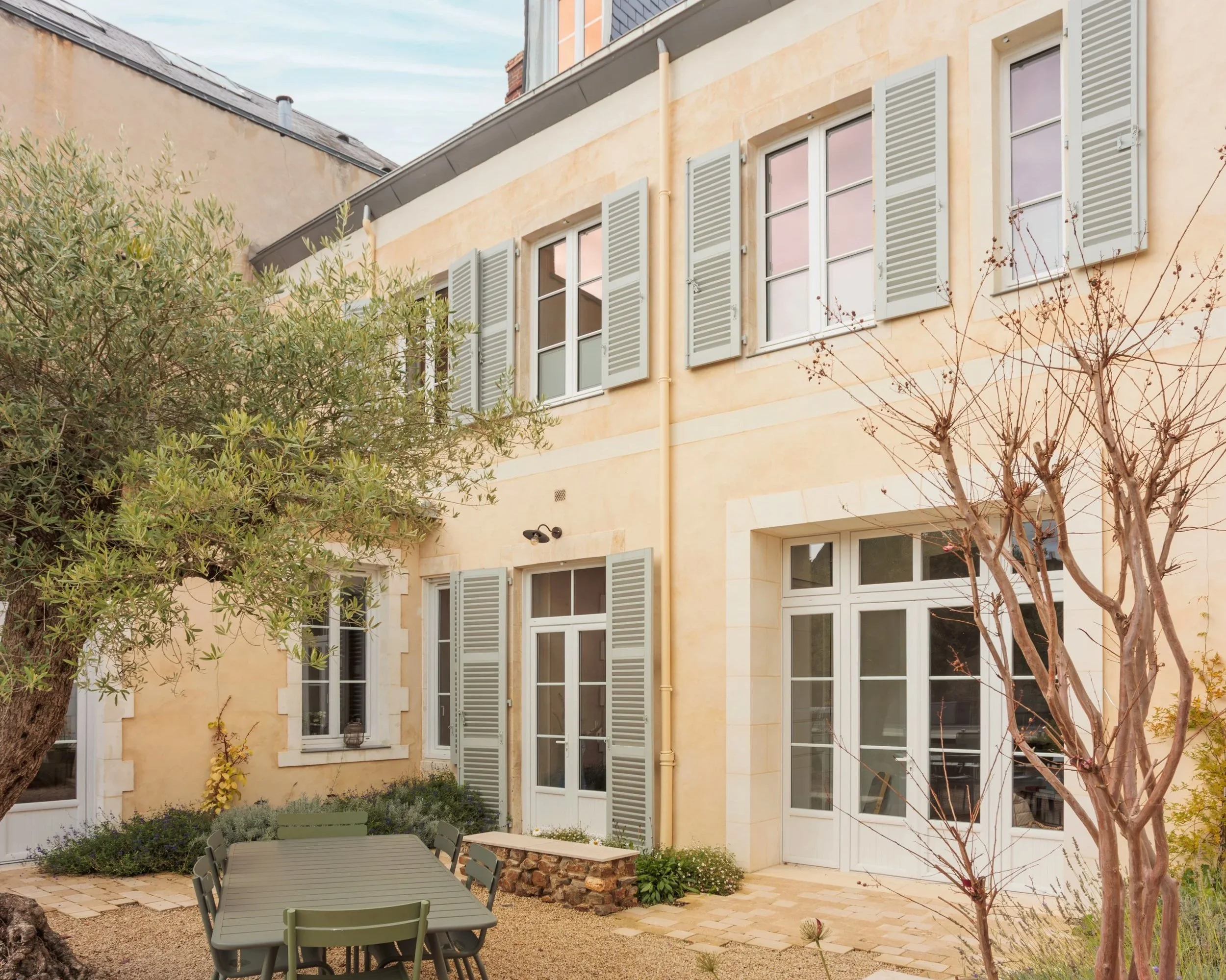Une cour intérieure avec une façade de bâtiment, plusieurs fenêtres avec volets gris, une table avec des chaises vertes, des arbres et des plantes décoratives.
