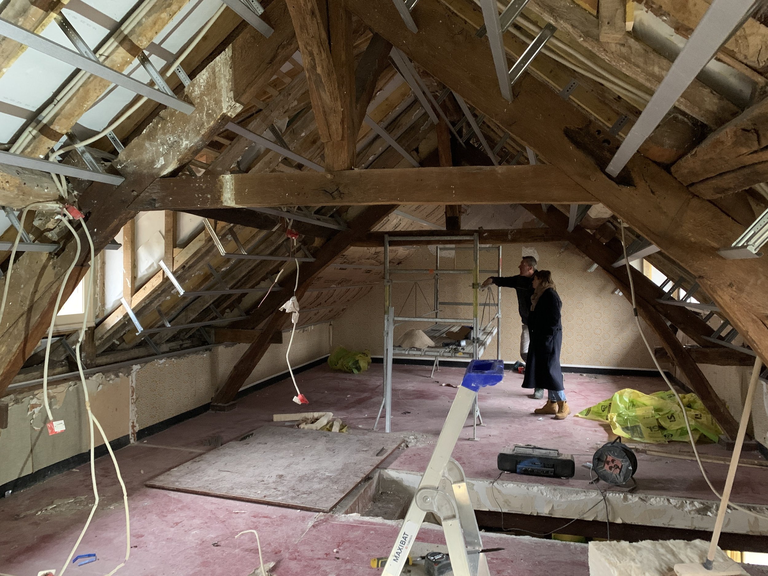 Deux personnes inspectant une pièce en rénovation ou en construction dans un grenier ou un loft, avec des éléments de structure en bois et des câbles électriques suspendus.