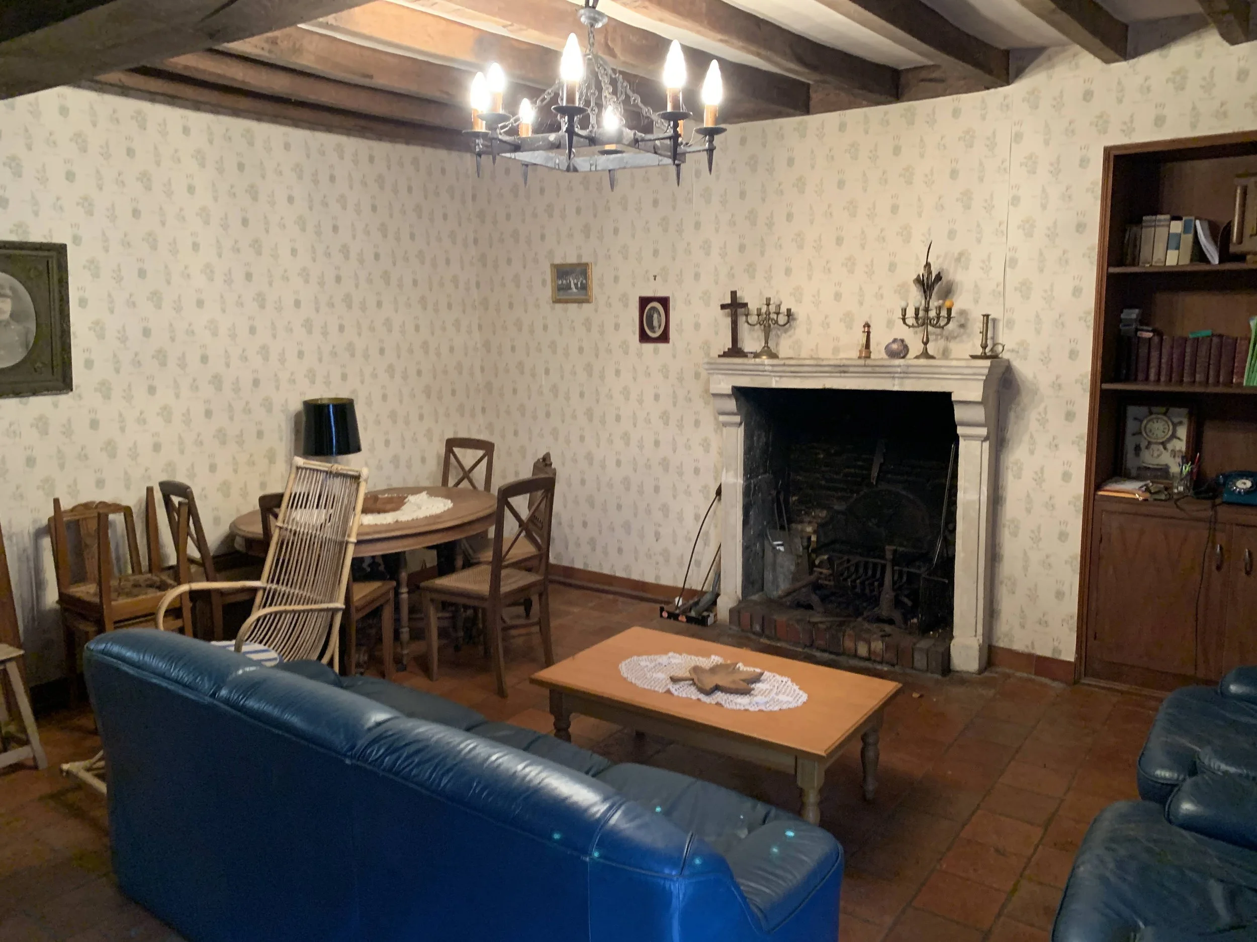 Avant la métamorphose - Salon avec canapé en cuir bleu, cheminée en marbre, table en bois, chaises en bois, décor vintage, lampadaire noir, chandelier suspendu, murs à papier peint, étagère en bois avec livres, bibelots, et téléphone ancien.