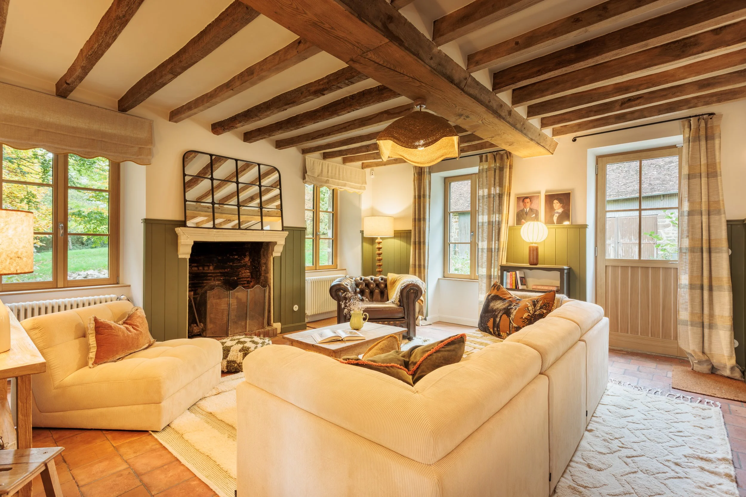 Salon de style rustique avec canapé beige, cheminée en pierre, cheminée en métal, deux fenêtres avec rideaux à carreaux, luminaire suspendu, lampes de table, tapis beige, meubles en bois, photos sur le mur, ambiance chaleureuse