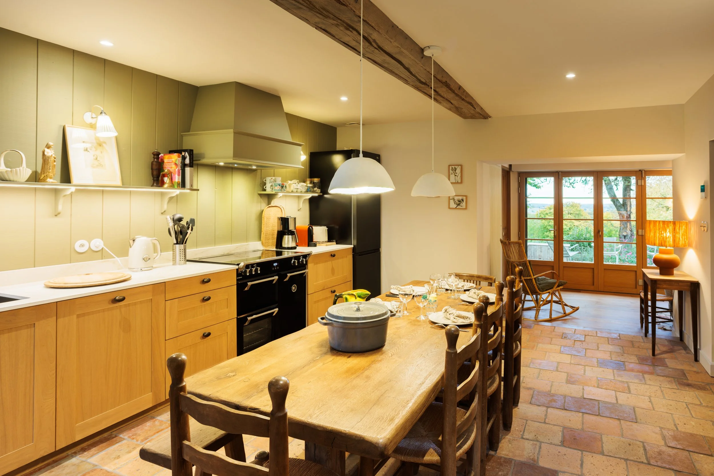 Cuisine chaleureuse avec plan de travail en bois, cuisine équipée d'un four, réfrigérateur noir, et des luminaires suspendus blancs. Une grande table en bois avec des assiettes, des verres et une cocotte en fonte. Au fond, une porte-fenêtre donnant sur un espace extérieur avec des arbres, et un coin salon avec un rocking-chair en bois, une lampe à côté et une petite table.