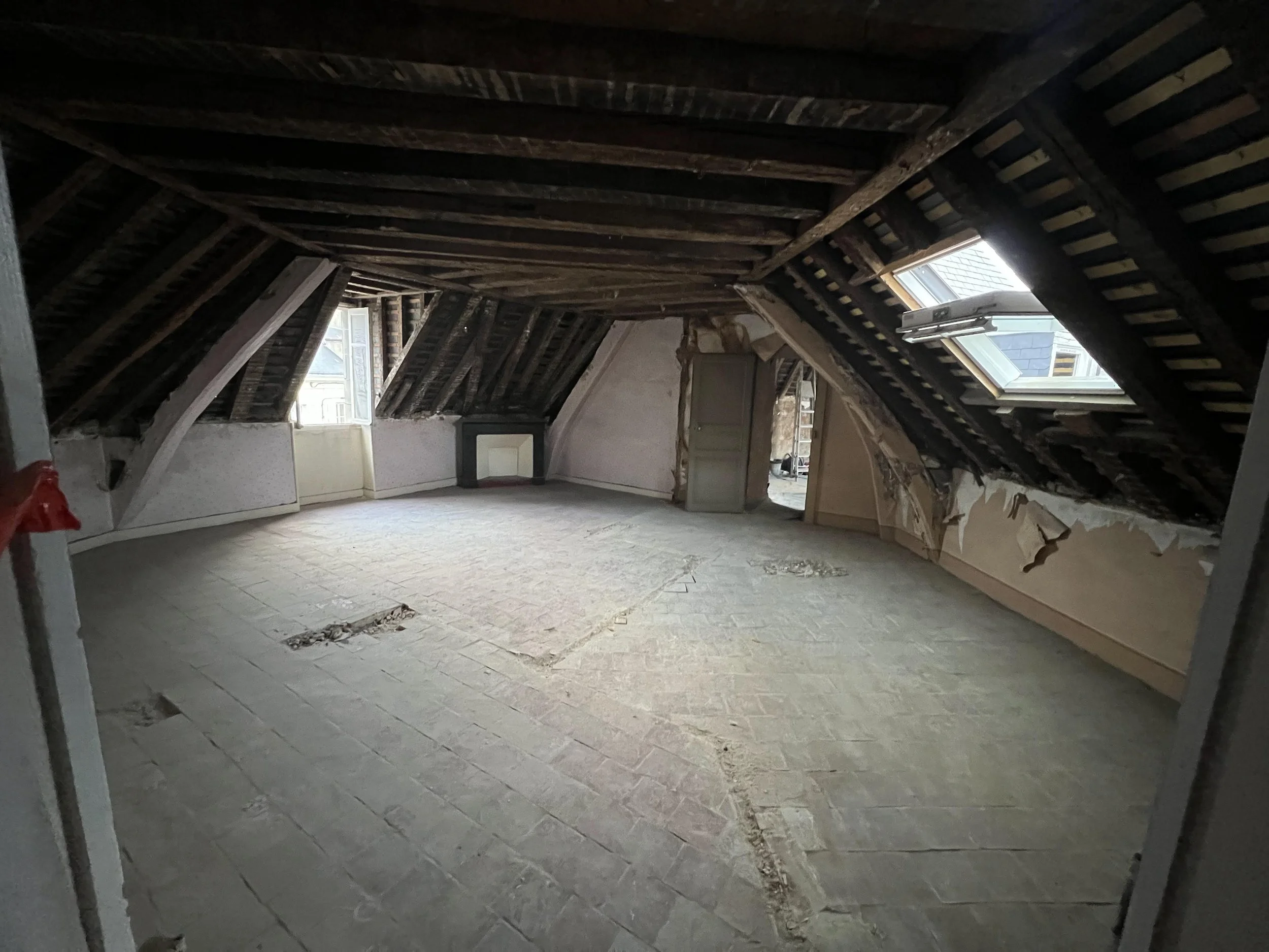 Une pièce en rénovation avec un plafond en pente, des fenêtres laissant entrer de la lumière, un sol en pierre, des murs partiellement endommagés et une porte au fond.