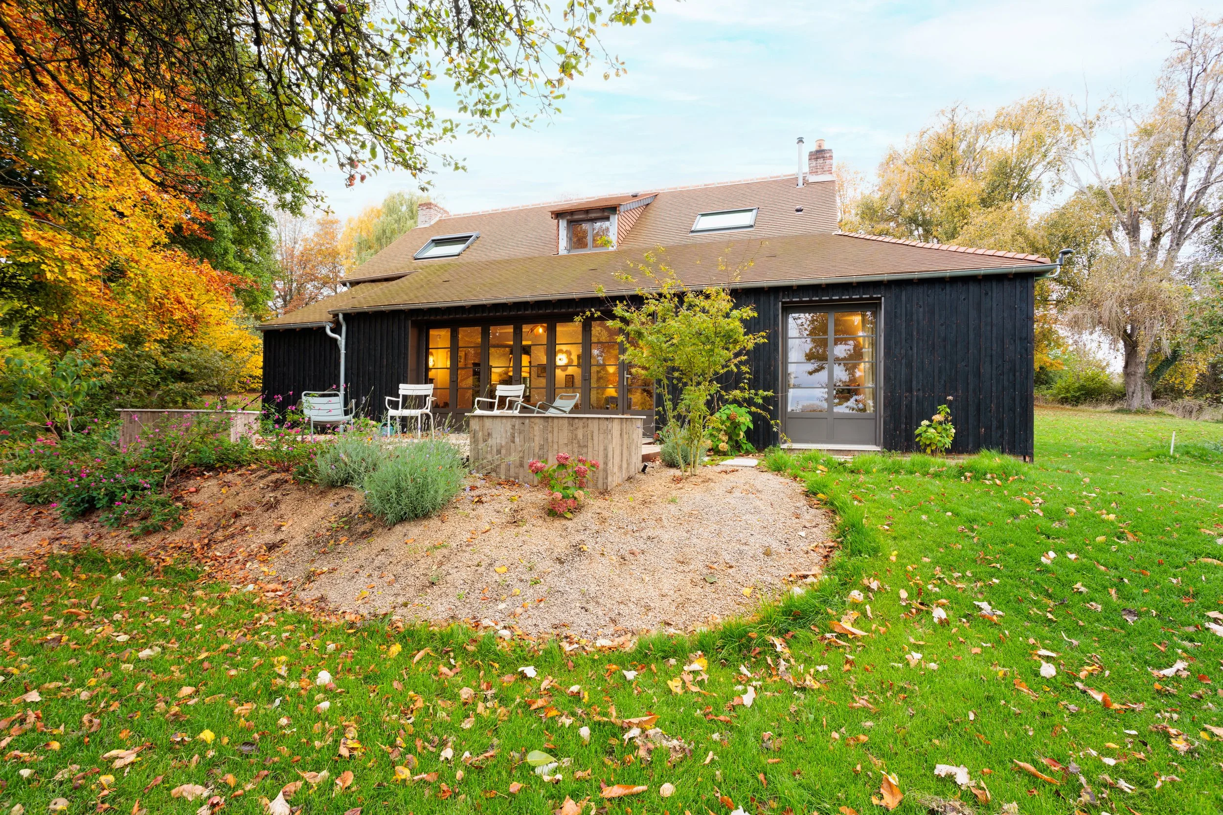 Maison en bois noir avec grandes fenêtres, terrasse avec mobilier d'extérieur, entourée de jardin et arbres en automne.