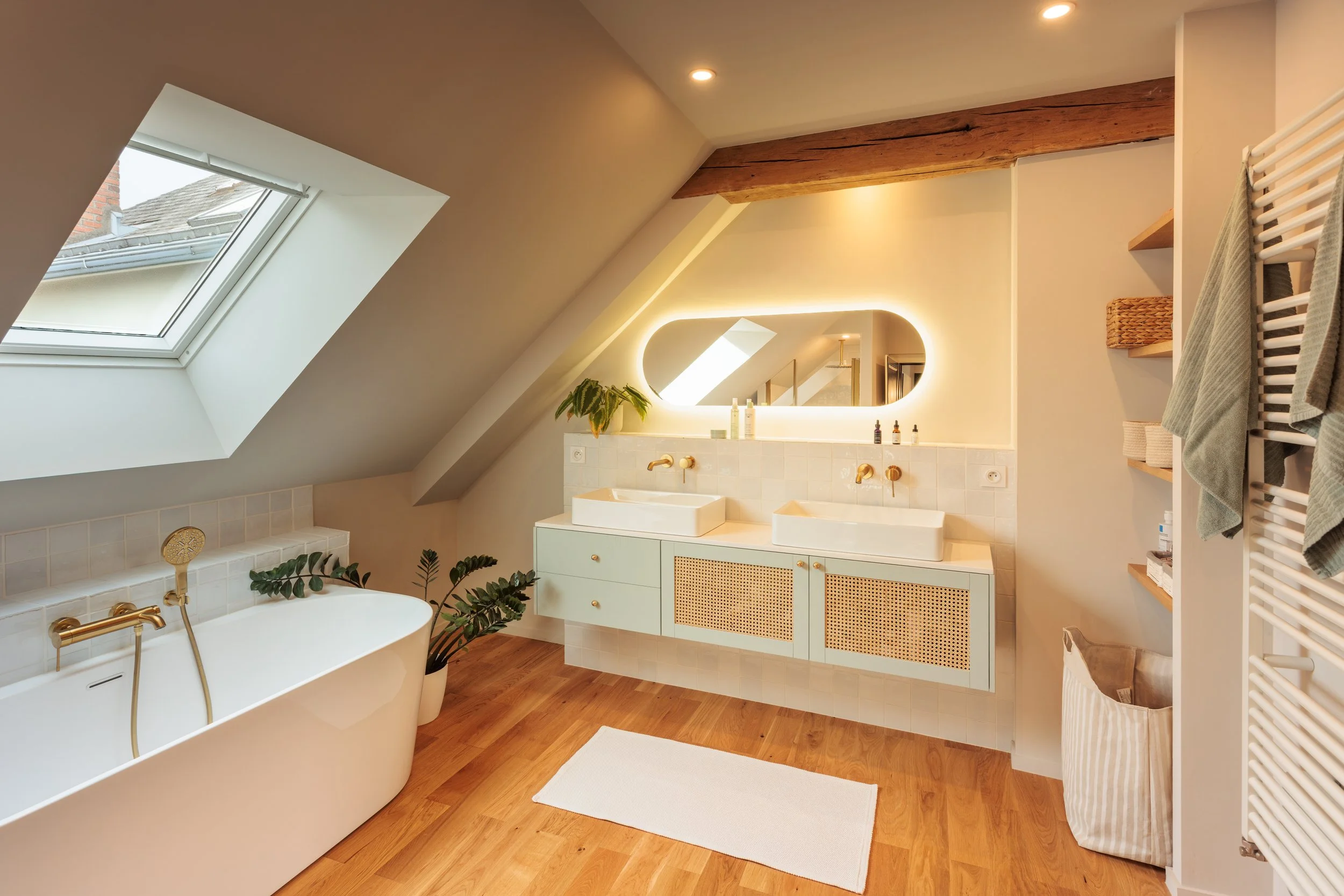 Salle de bain lumineuse avec baignoire, lavabo double, grand miroir à éclairage intégré, fenêtre de toit, étagères en bois, plantes vertes, tapis blanc, serviettes, sol en bois.