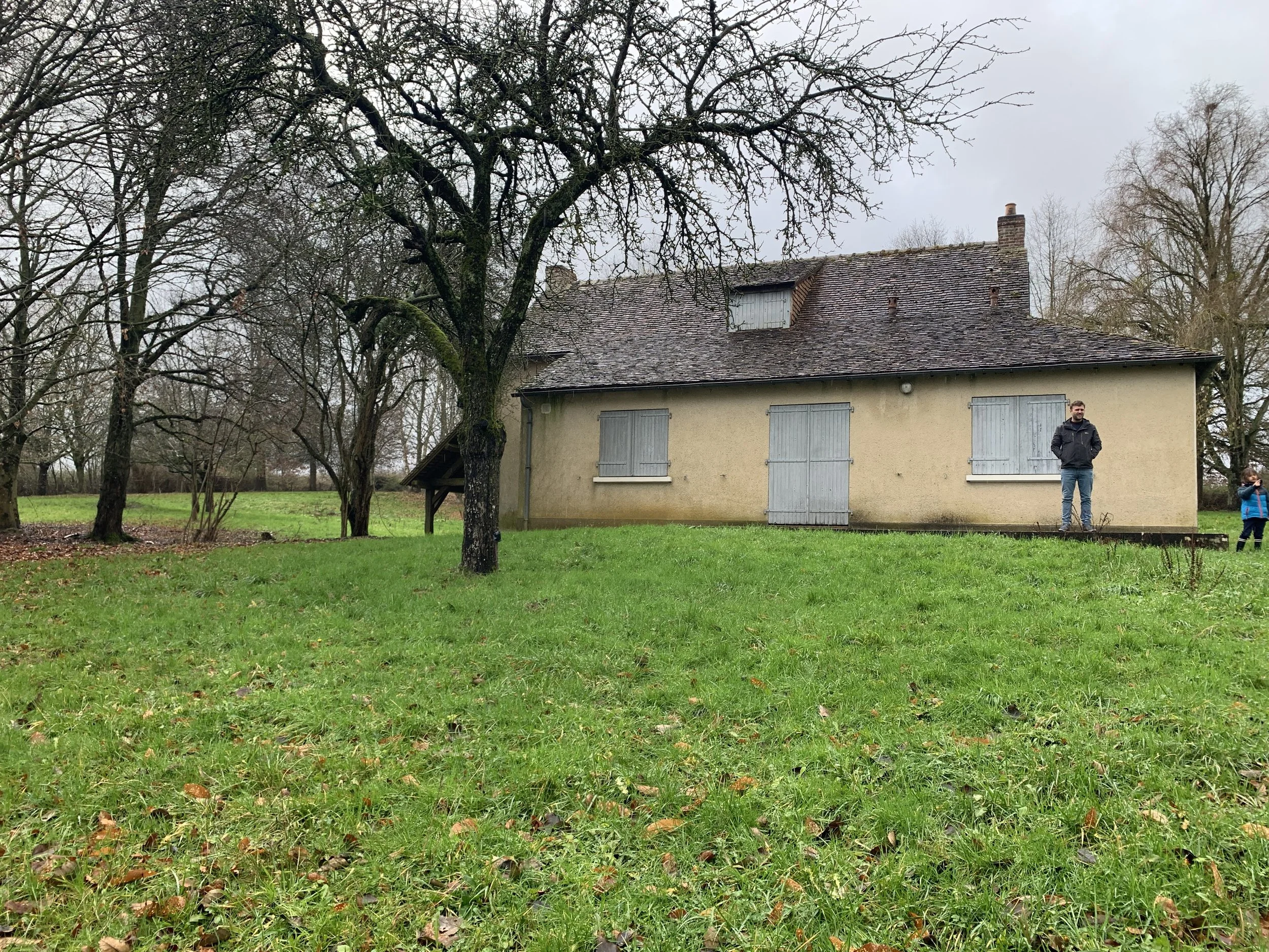 Une maison ancienne avec des volets en bois gris, entourée d'arbres nus en hiver, dans le jardin verdoyant.