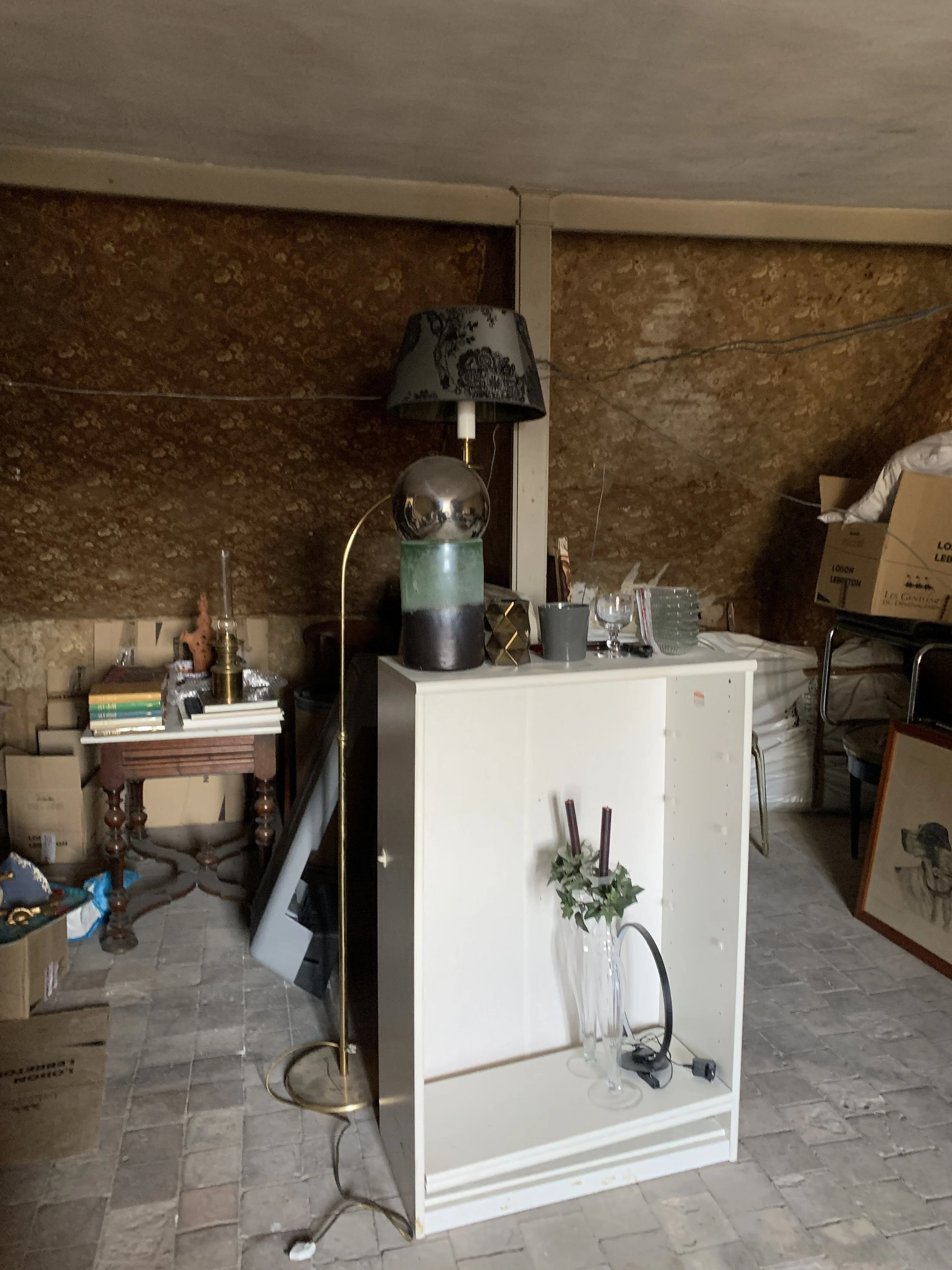 Une pièce sous pente en désordre avec des meubles et objets décoratifs divers, des livres, des boîtes et des cadres empilés, avant la transformation