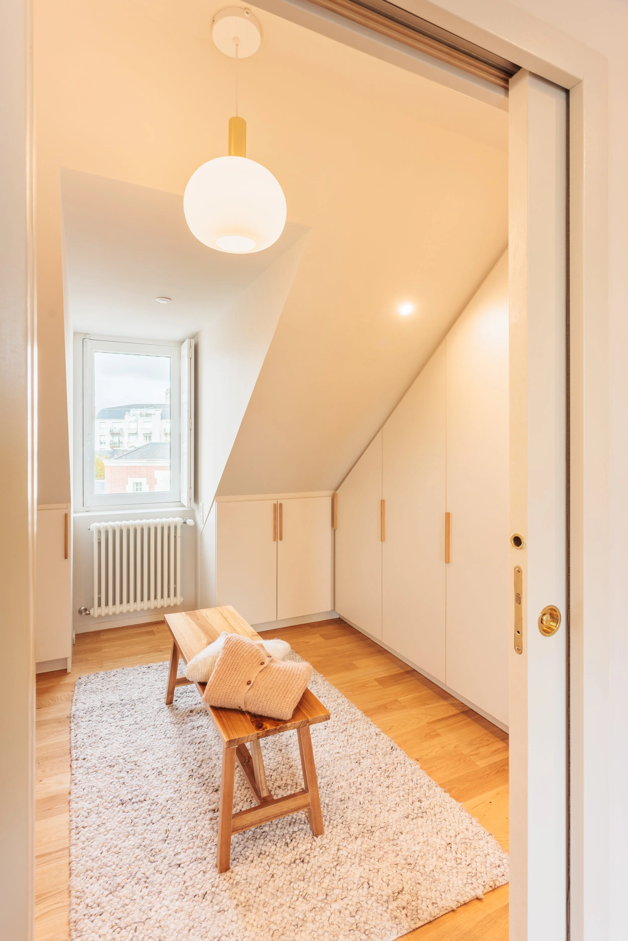 Petite pièce avec un sol en bois, un tapis beige, des armoires blanches, une fenêtre avec vue sur des bâtiments, un radiateur, un plafonnier arrondi, une banquette en bois avec un pull beige dessus