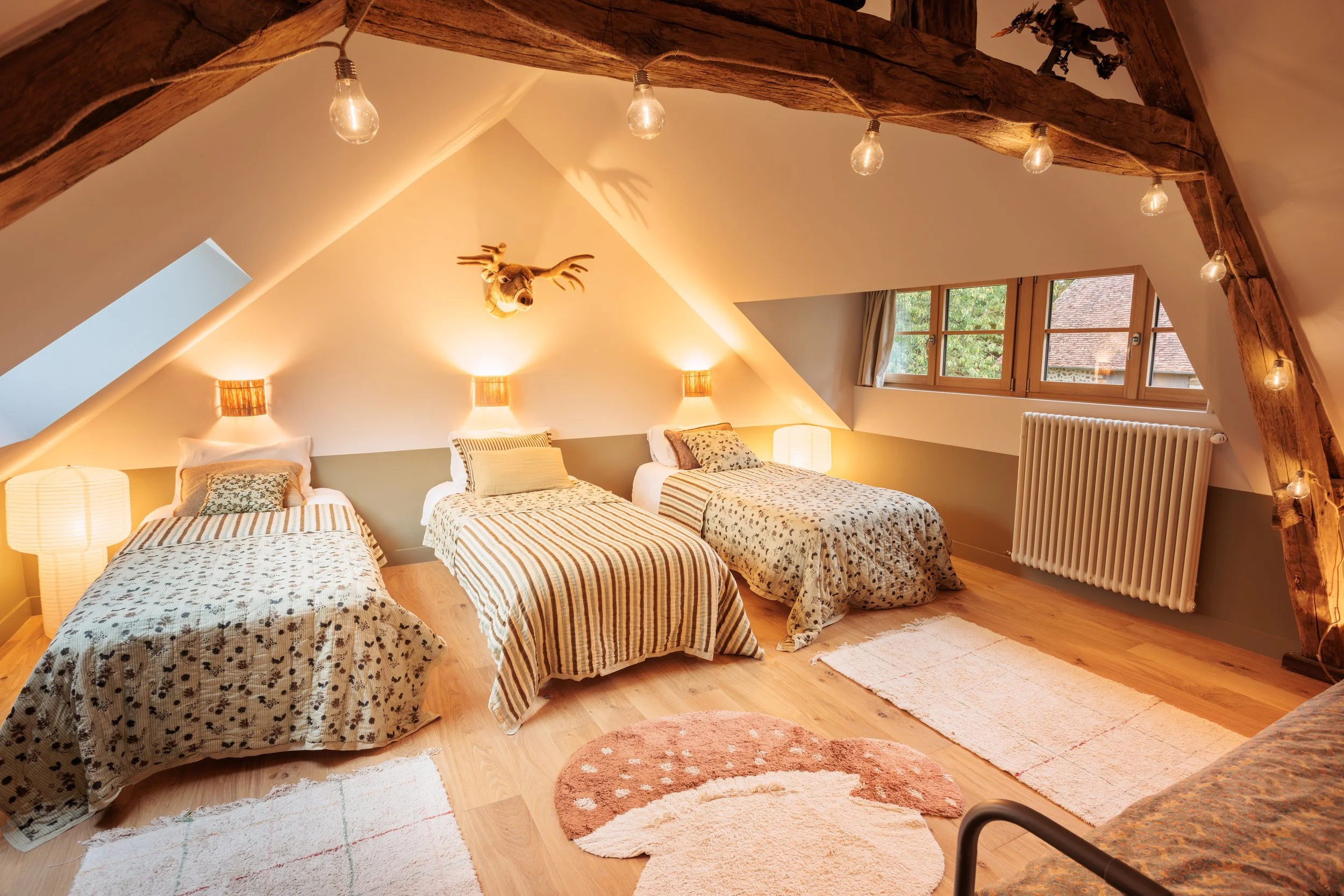 Chambre d'auberge avec trois lits, décoration rustique, murs peints en deux tons avec éclairage chaud, fenêtres en bois, décoration de trophée de cerf, tapis moelleux, éclairage par lampes de chevet, ambiance chaleureuse.