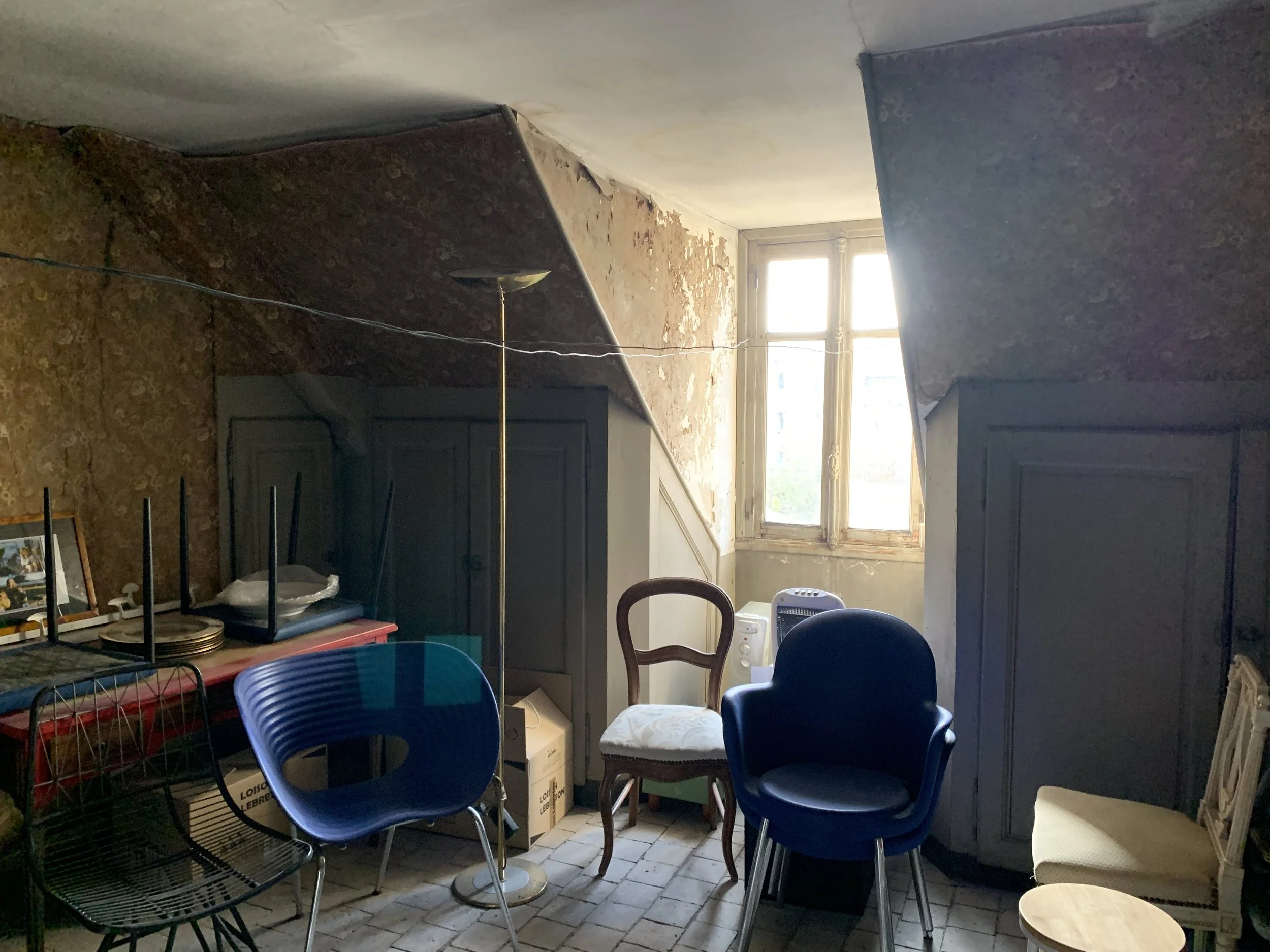 Avant la rénovation, pièce sous combles, encombrée de meubles simples, une fenêtre qui laisse passer la lumière, sol en briques