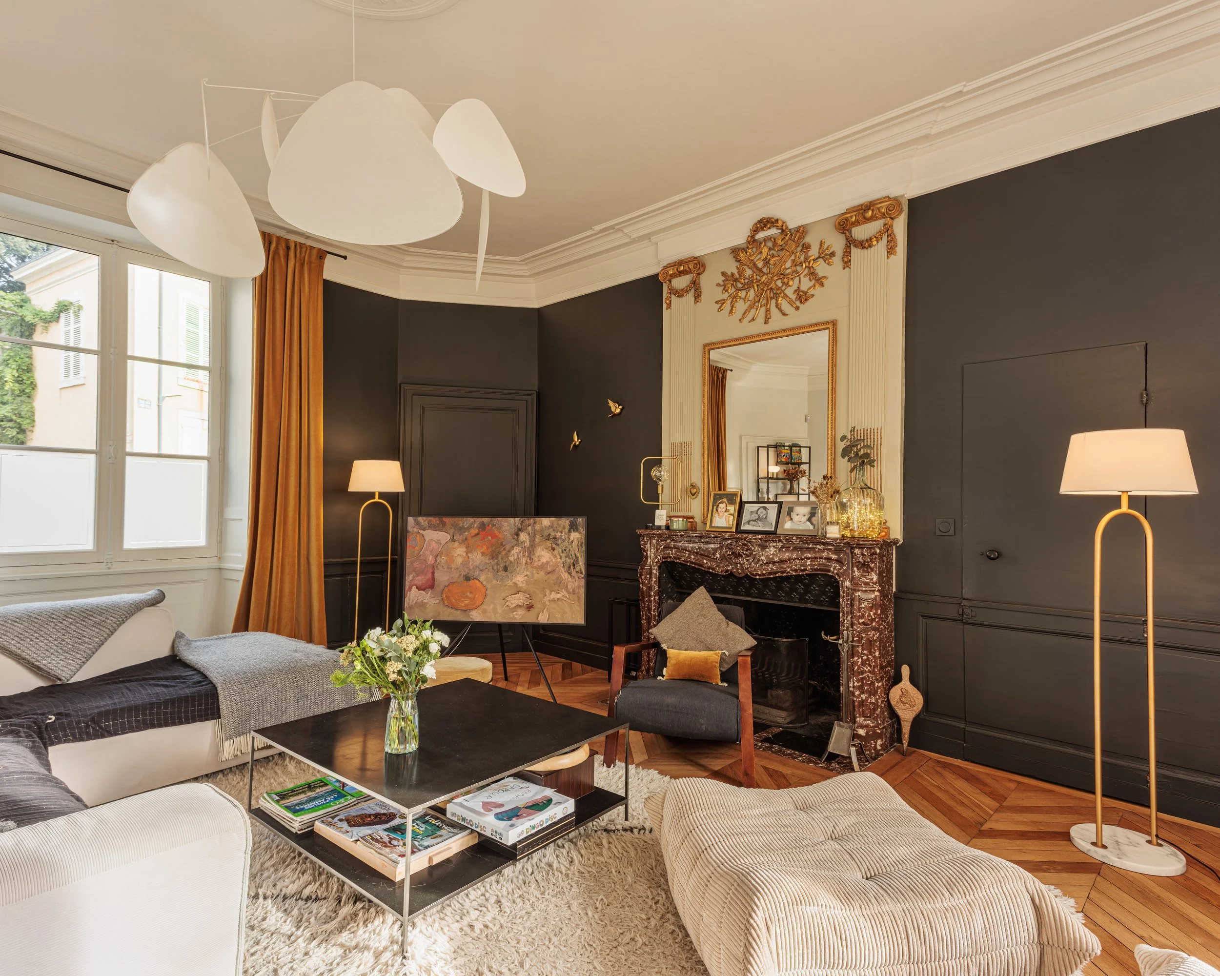 Salon de style classique avec cheminée en marbre, cheminée ornée de photos et objets décoratifs, grand miroir doré, fauteuils et canapé, tables basses, lampes sur pied, grandes fenêtres avec rideaux dorés, plafond décoré.
