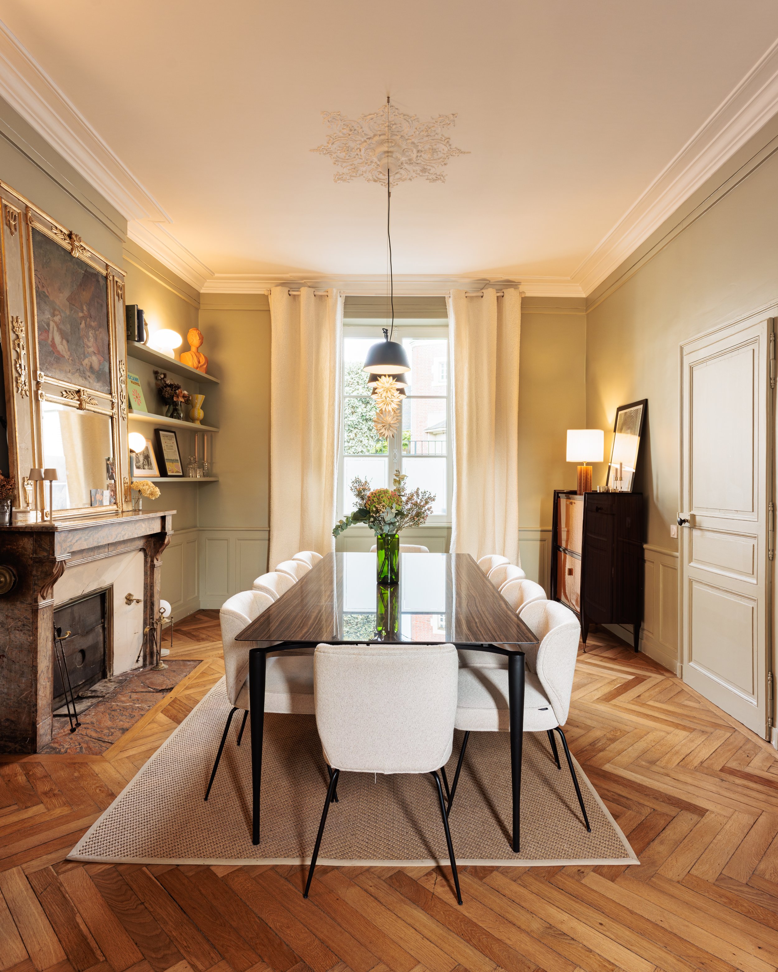 Salle à manger élégante avec un grand tableau, cheminée en marbre, et un grand tapis beige sous une table en bois avec neuf chaises blanches entourant une vase de fleurs au centre, avec des rideaux blancs, un lustre, et un meuble en bois avec une lampe dans la pièce.
