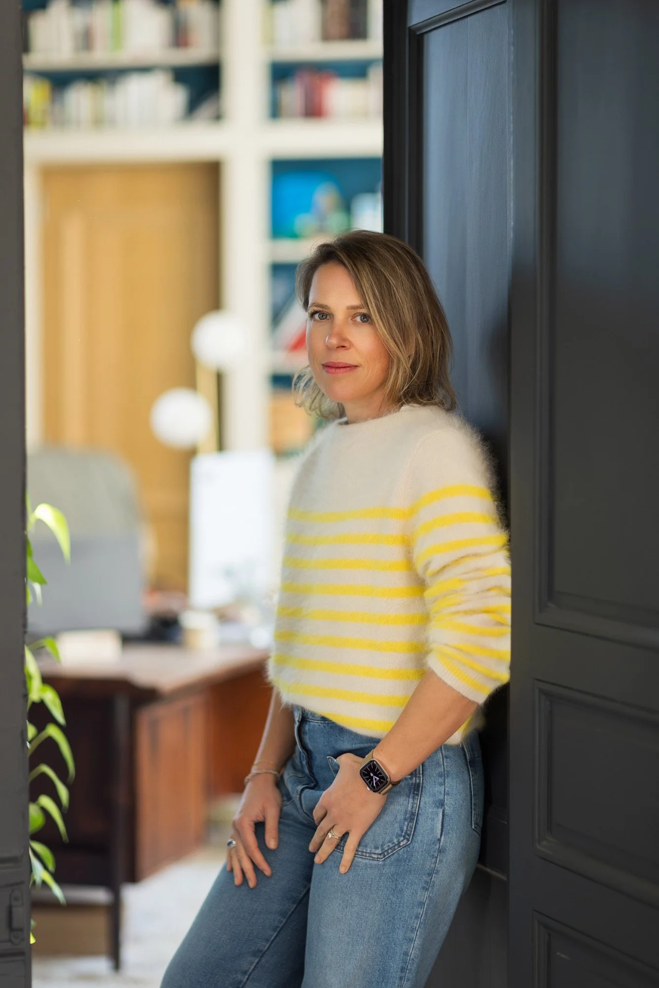 Femme debout dans une pièce lumineuse, portant un pull rayé jaune et blanc, et un jean, posant près d'une porte en bois bleu, avec une étagère remplie de livres en arrière-plan.