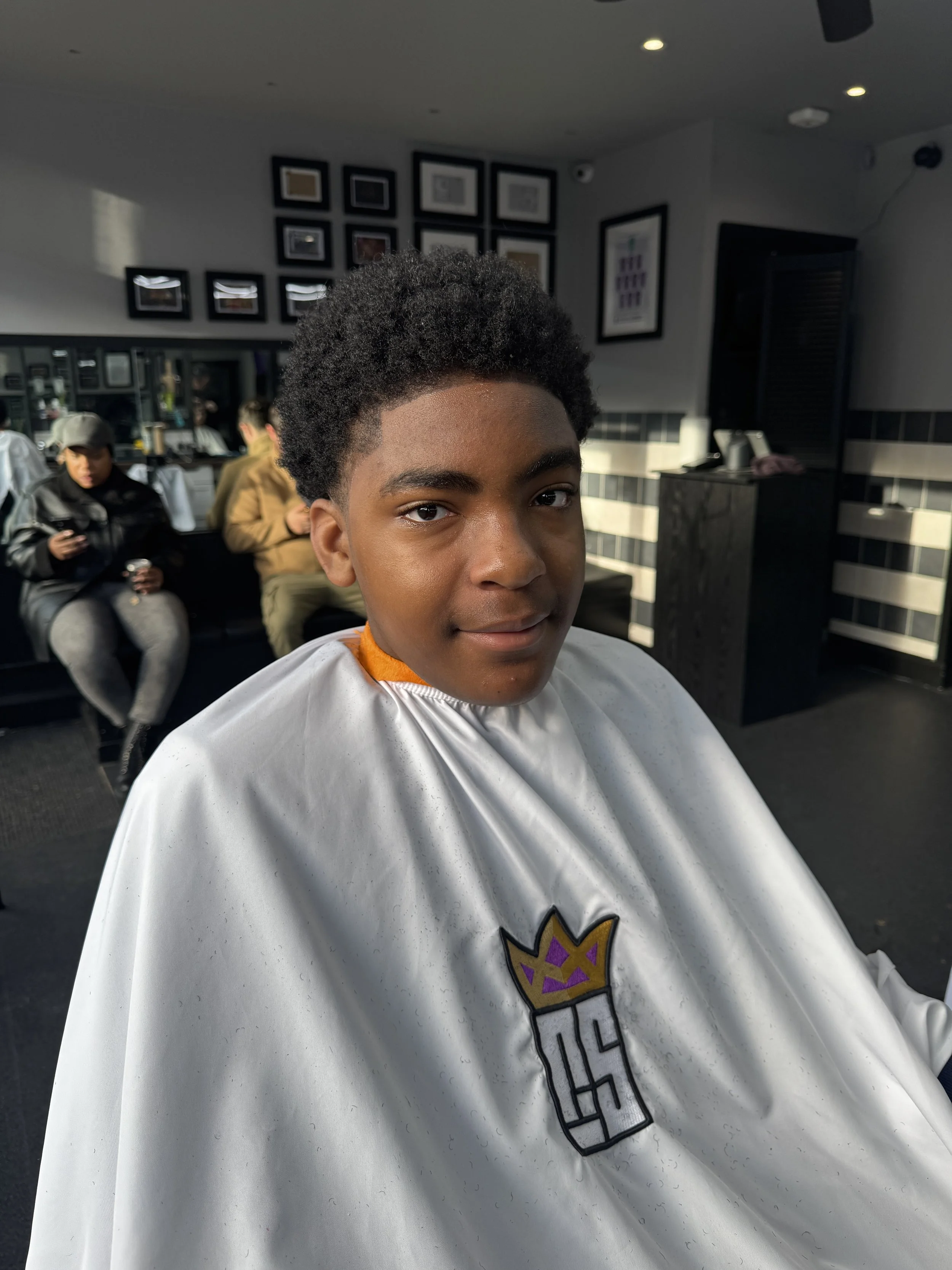 Young boy at a barbershop getting a haircut, wearing a white cape with a purple and gold crown logo.