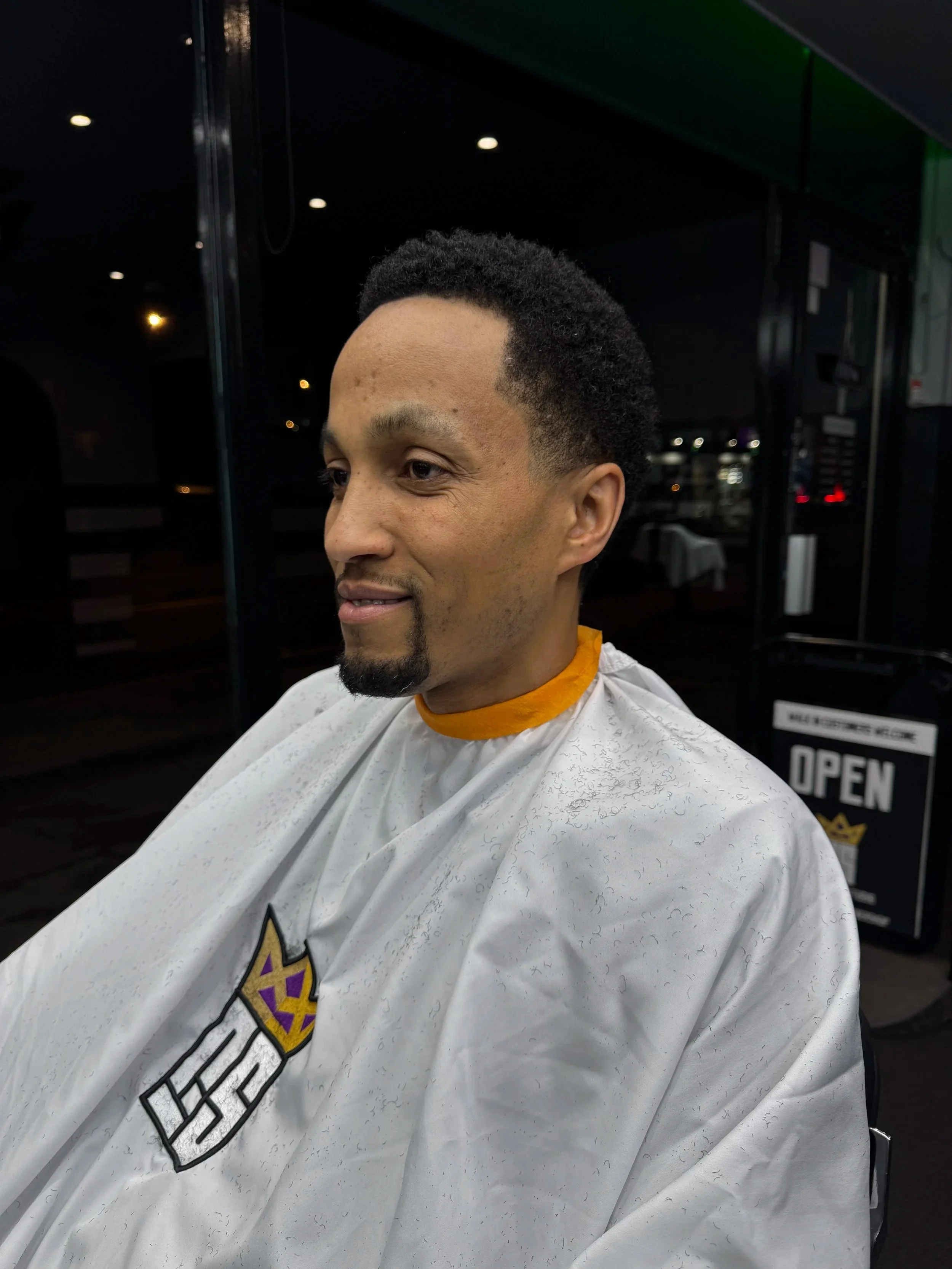 Man with curly black hair, goatee, and light brown skin, sitting in a salon chair, wearing a white barber cape with a logo.