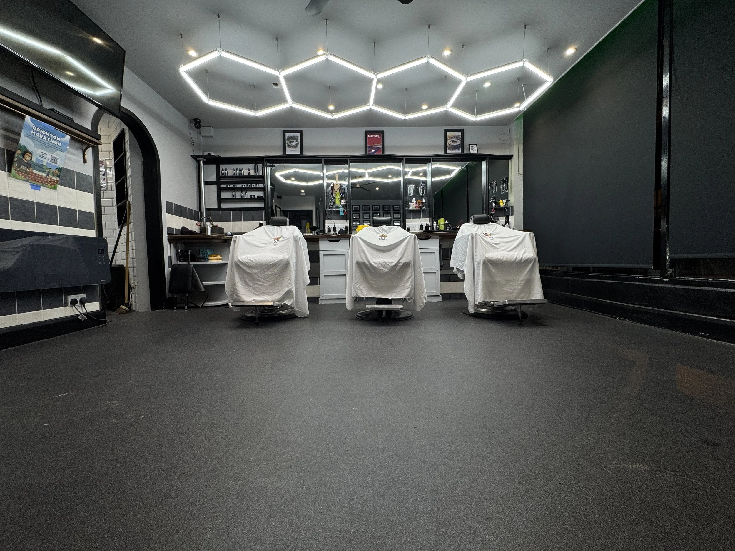 Three barber chairs with white capes in a barber shop, mirrored wall, modern hexagon light fixtures on the ceiling, black and white tiled walls, and various barber tools on shelves.