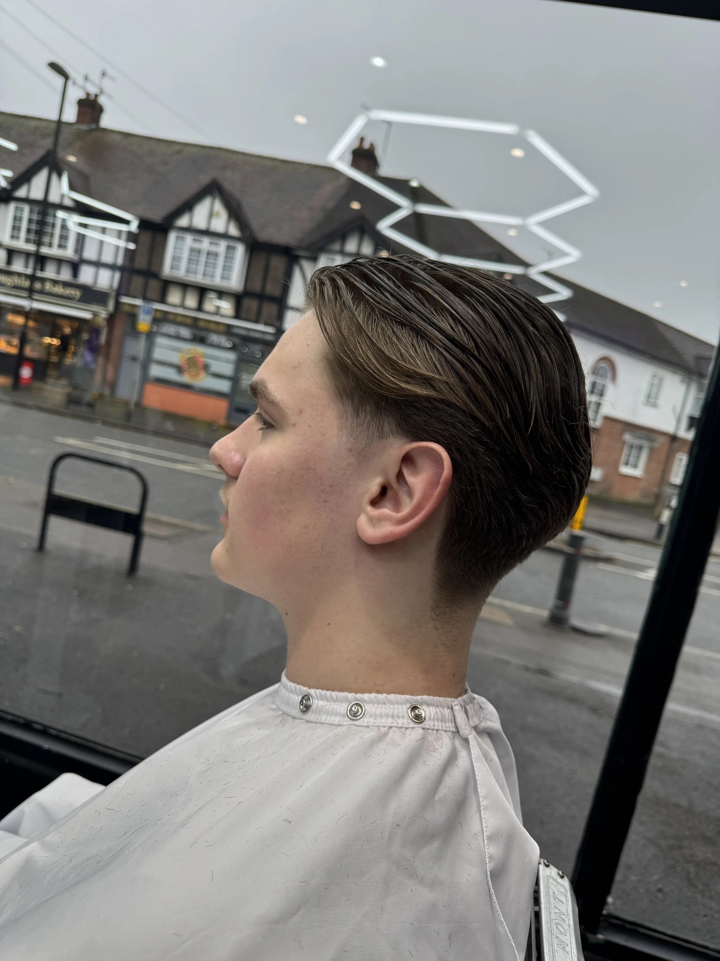 Side profile of a young man with neatly styled brown hair, sitting inside a vehicle and looking outside through a large window, with a view of houses and a street outside.