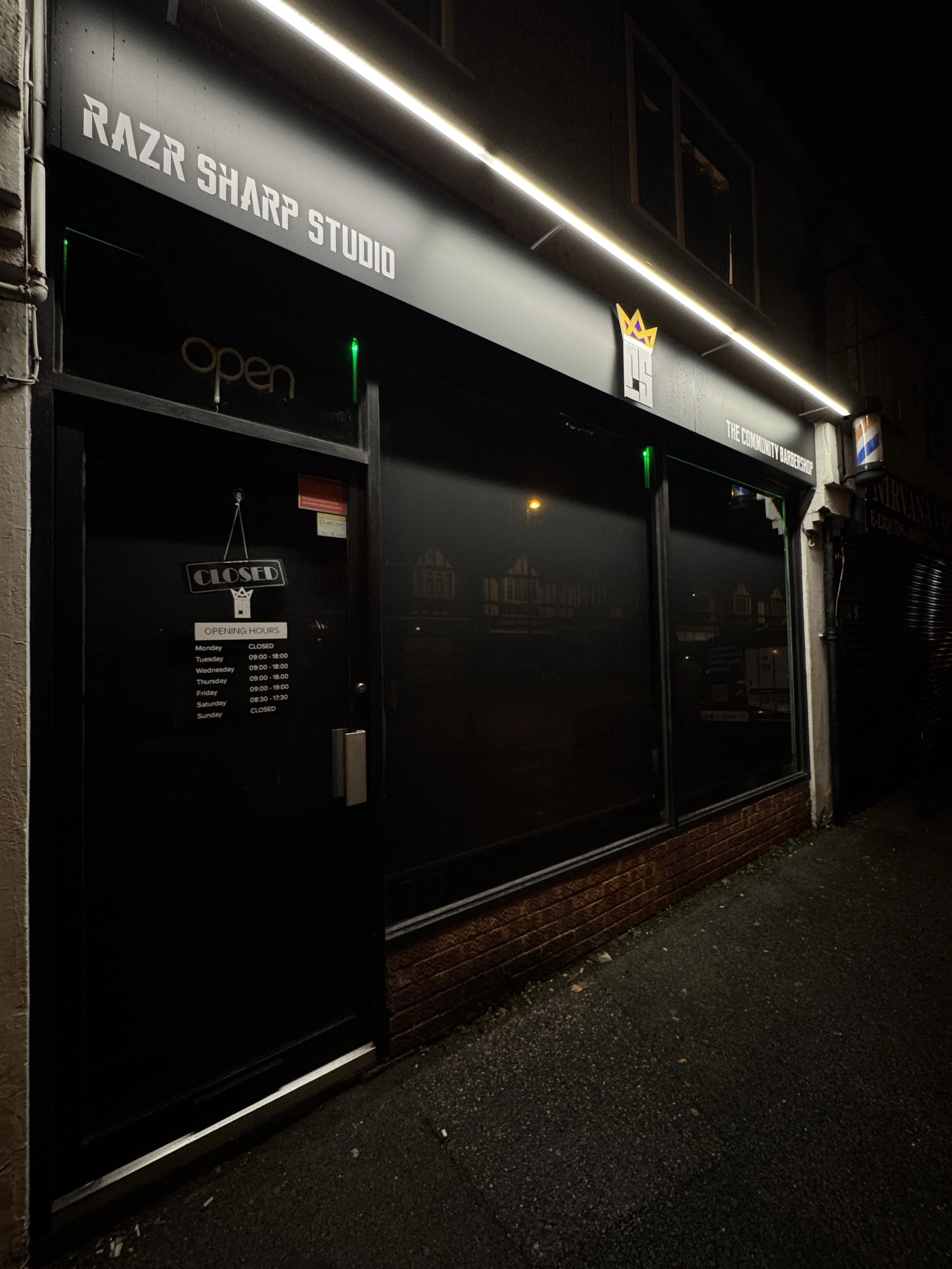 Black storefront with signs indicating it is open late at night, called RAZR SHARP STUDIO, with a door displaying the hours and a closed sign, and the sidewalk in front of it.