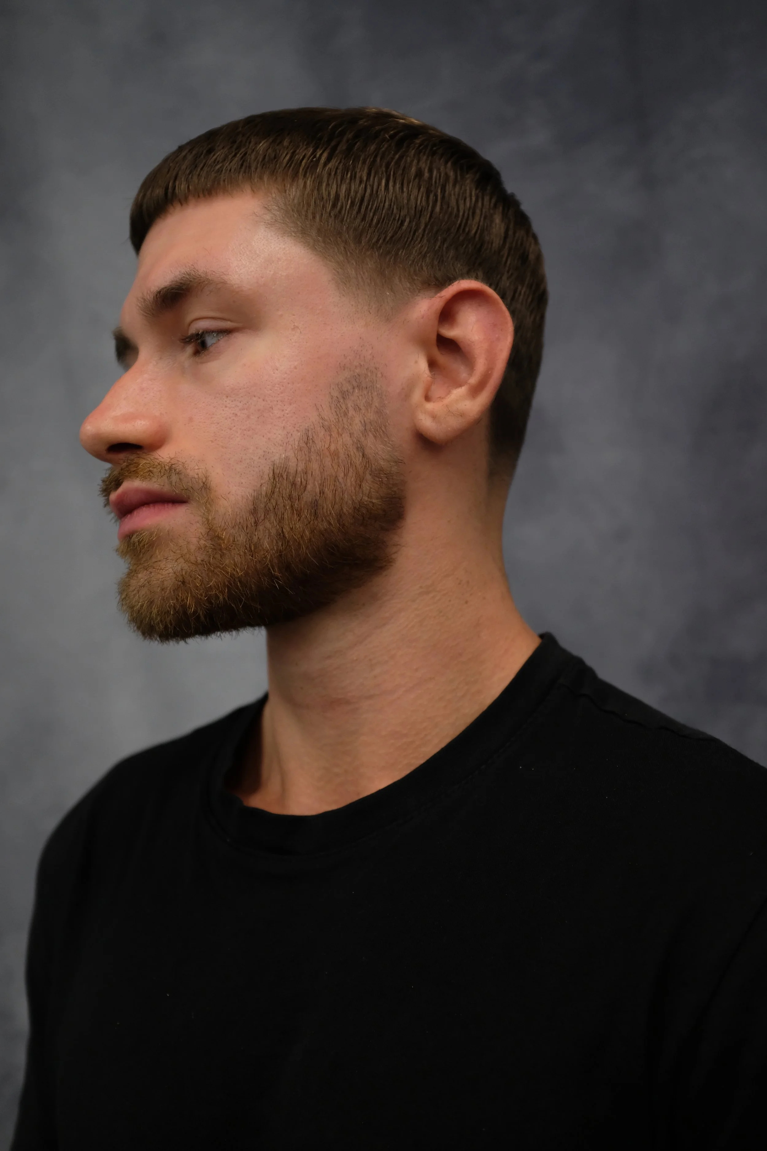 Side profile of a young man with short brown hair and a beard, wearing a black shirt, against a dark background.
