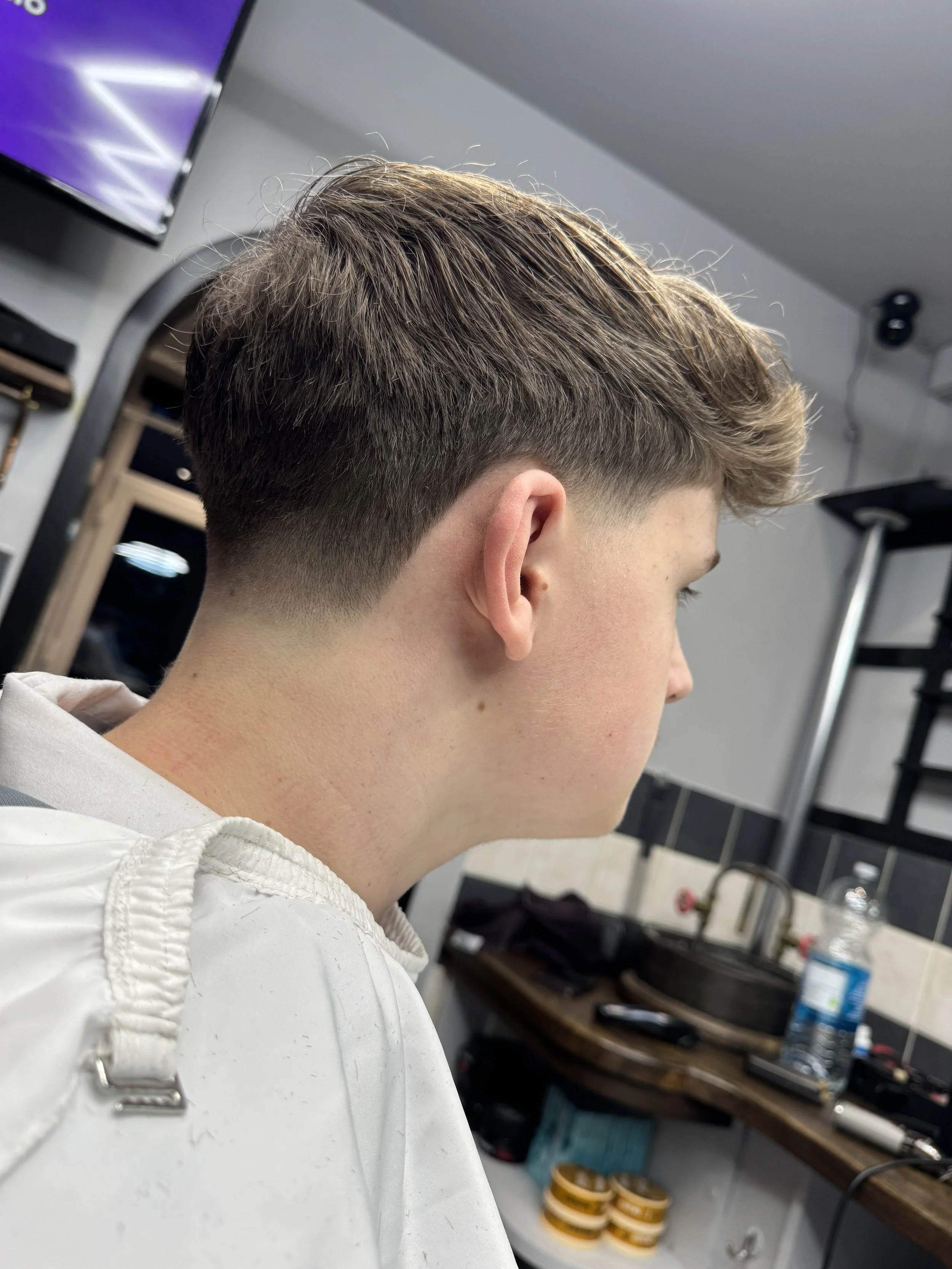 Side view of a young man with a fresh haircut, sitting in a barbershop with hair clippers nearby, reflected in a mirror.