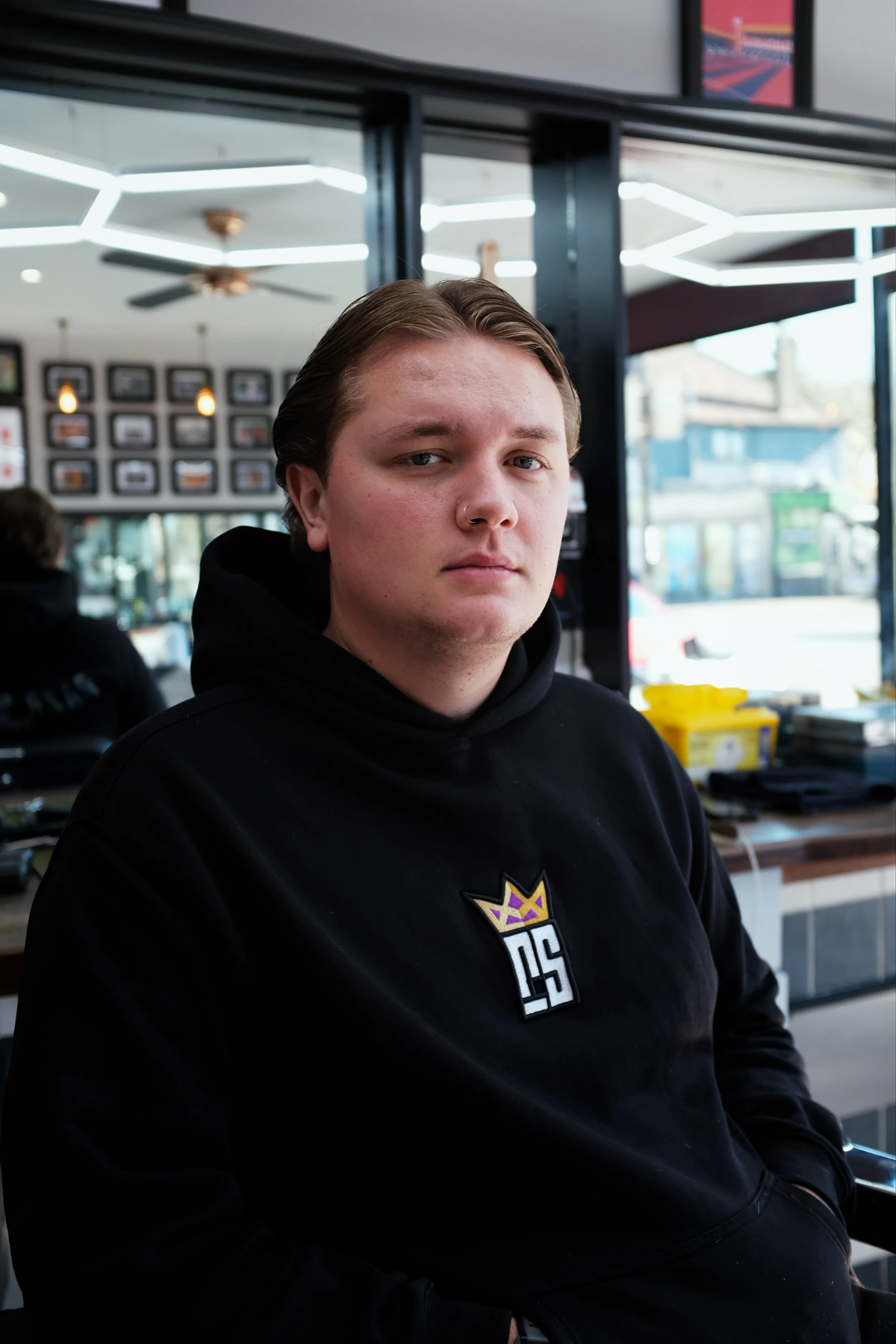 Young man with blonde hair and nose piercing sitting in a barbershop or salon with framed pictures on the wall, shelves with styling products, and mirrors behind him.