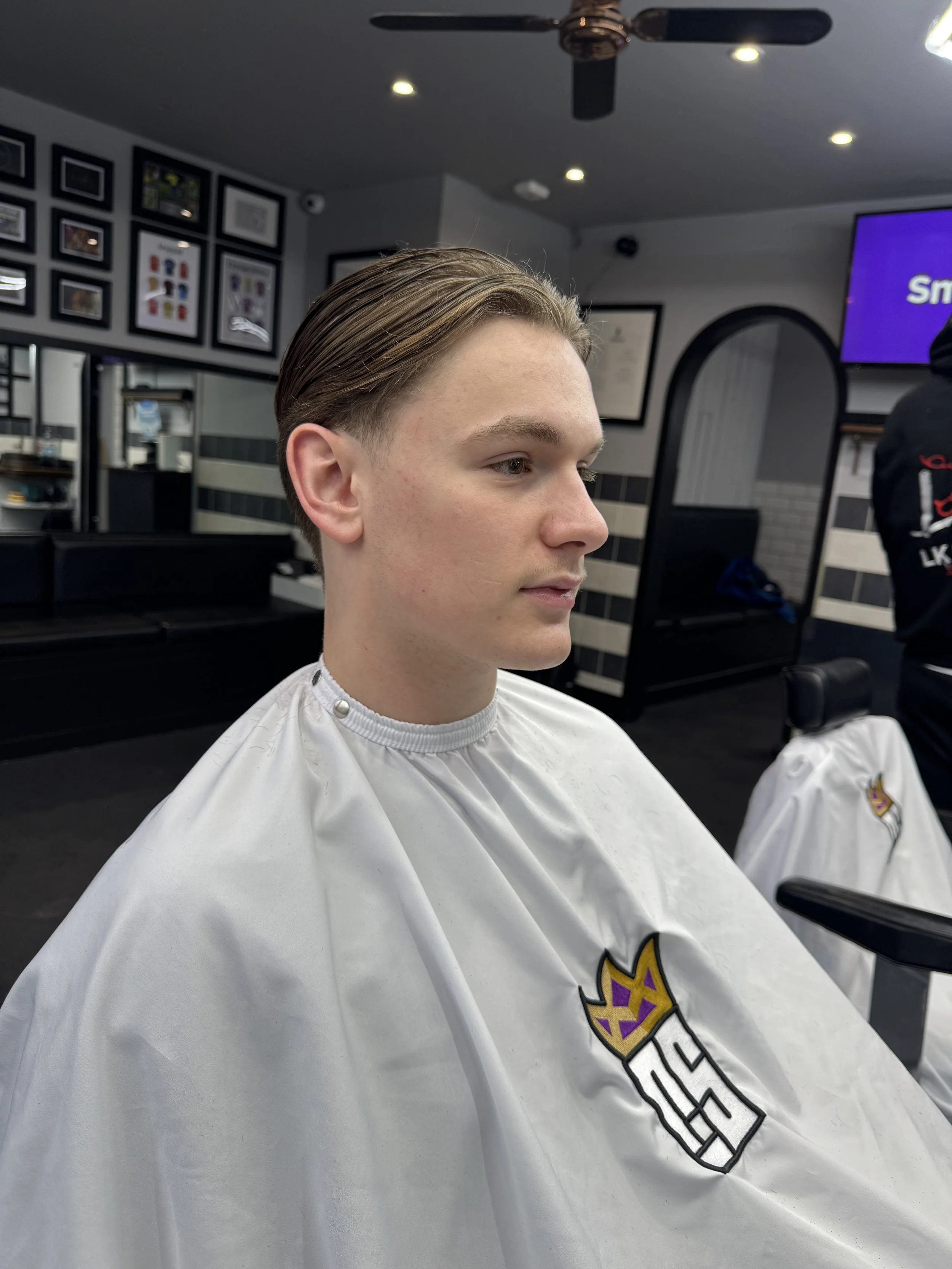 A young man with neatly combed hair getting a haircut at a barbershop, sitting in a chair covered with a white cape that has a graphic logo.