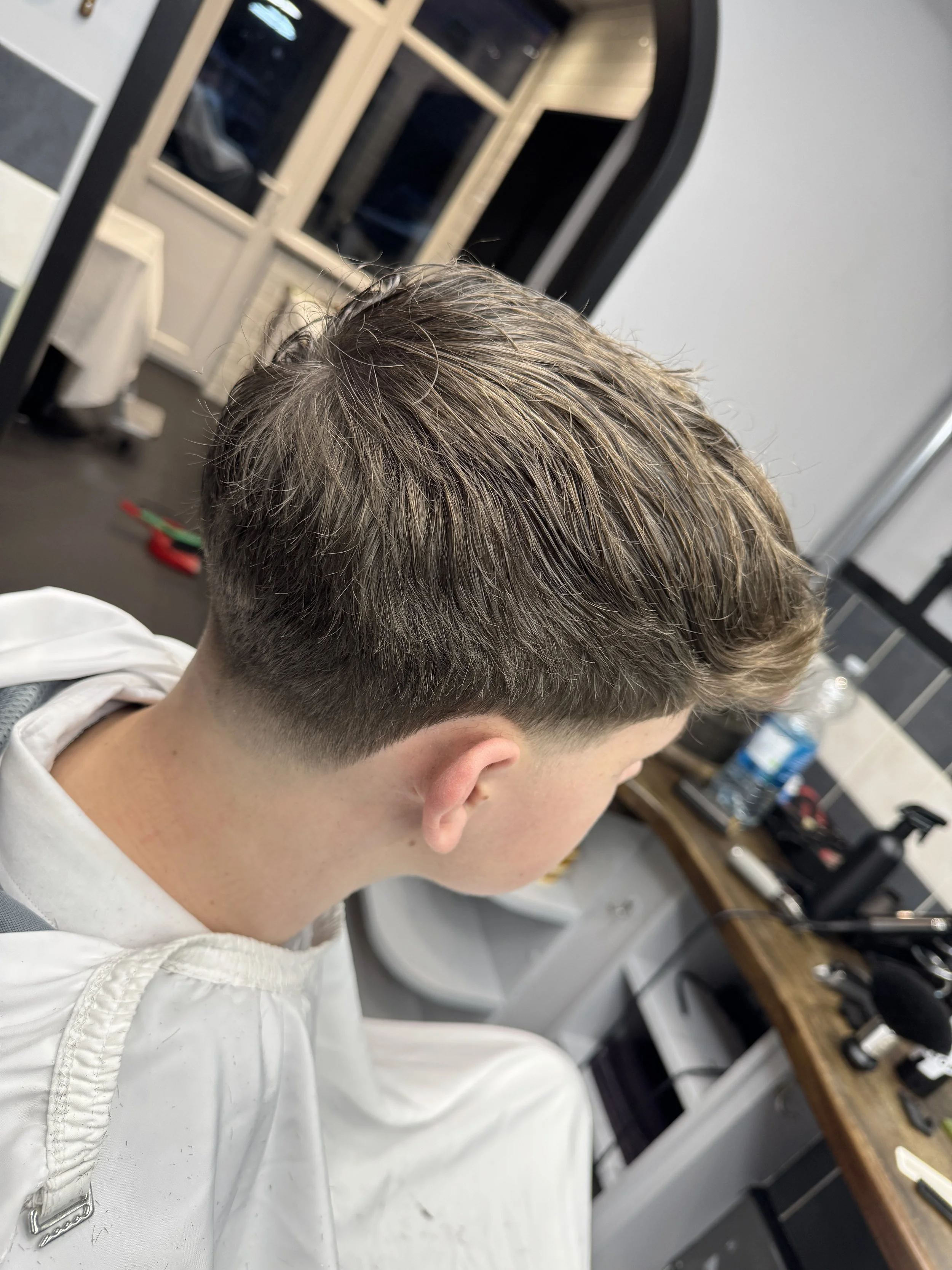 Young man with a fade haircut sitting in a barber shop, with barber tools on a wooden counter and a mirror in the background.